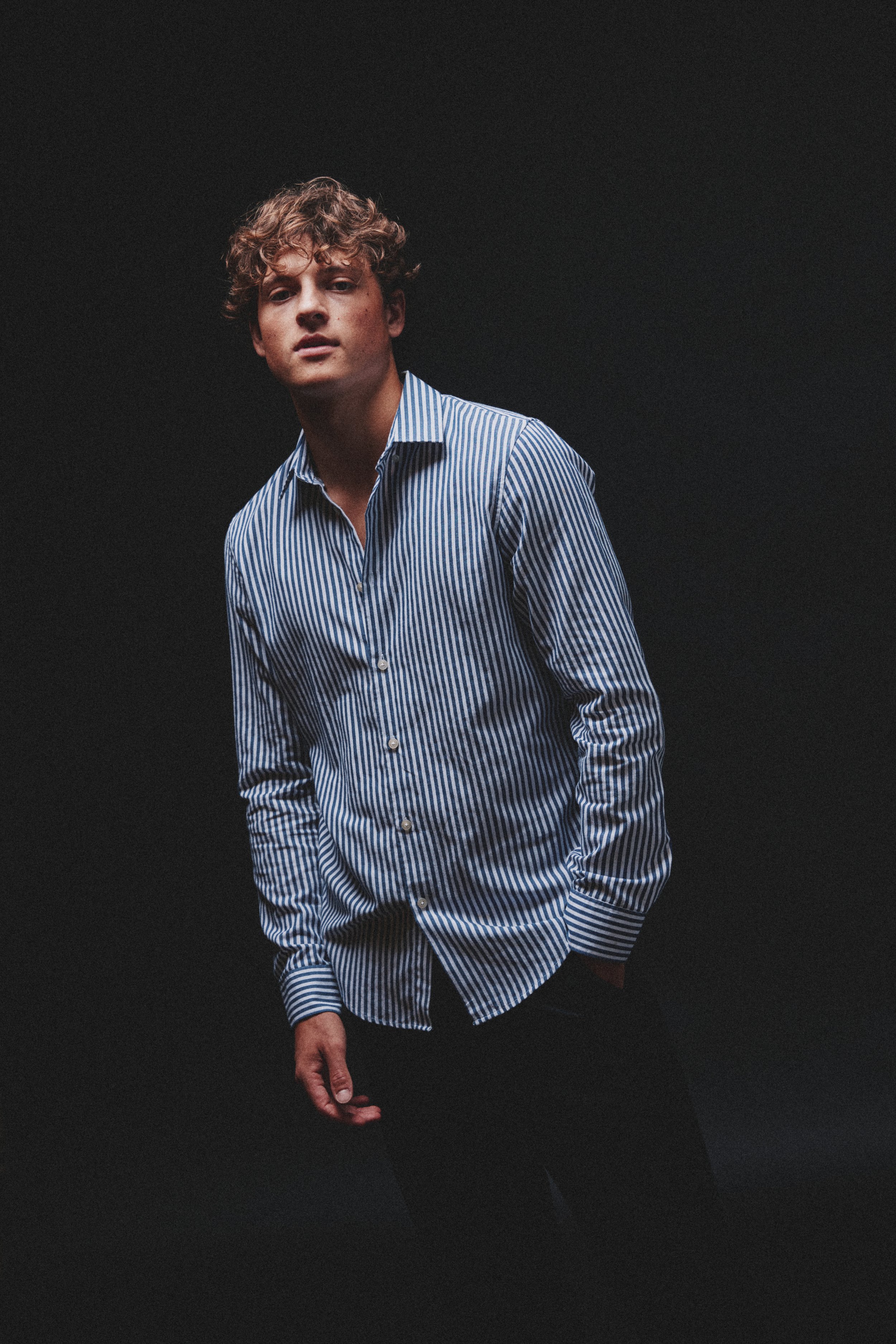 Portrait of a young man with curly hair wearing a blue and white striped button-up shirt standing against a dark background.