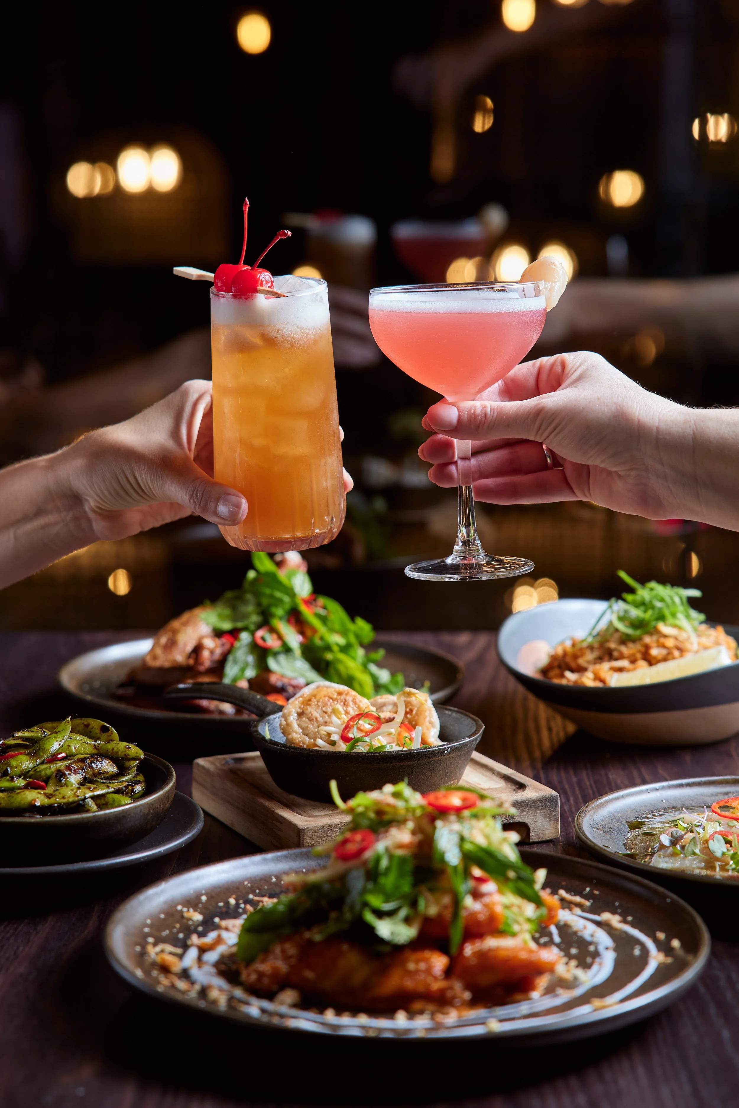 Two hands holding cocktails in front of a variety of Asian dishes on a dark wooden table.