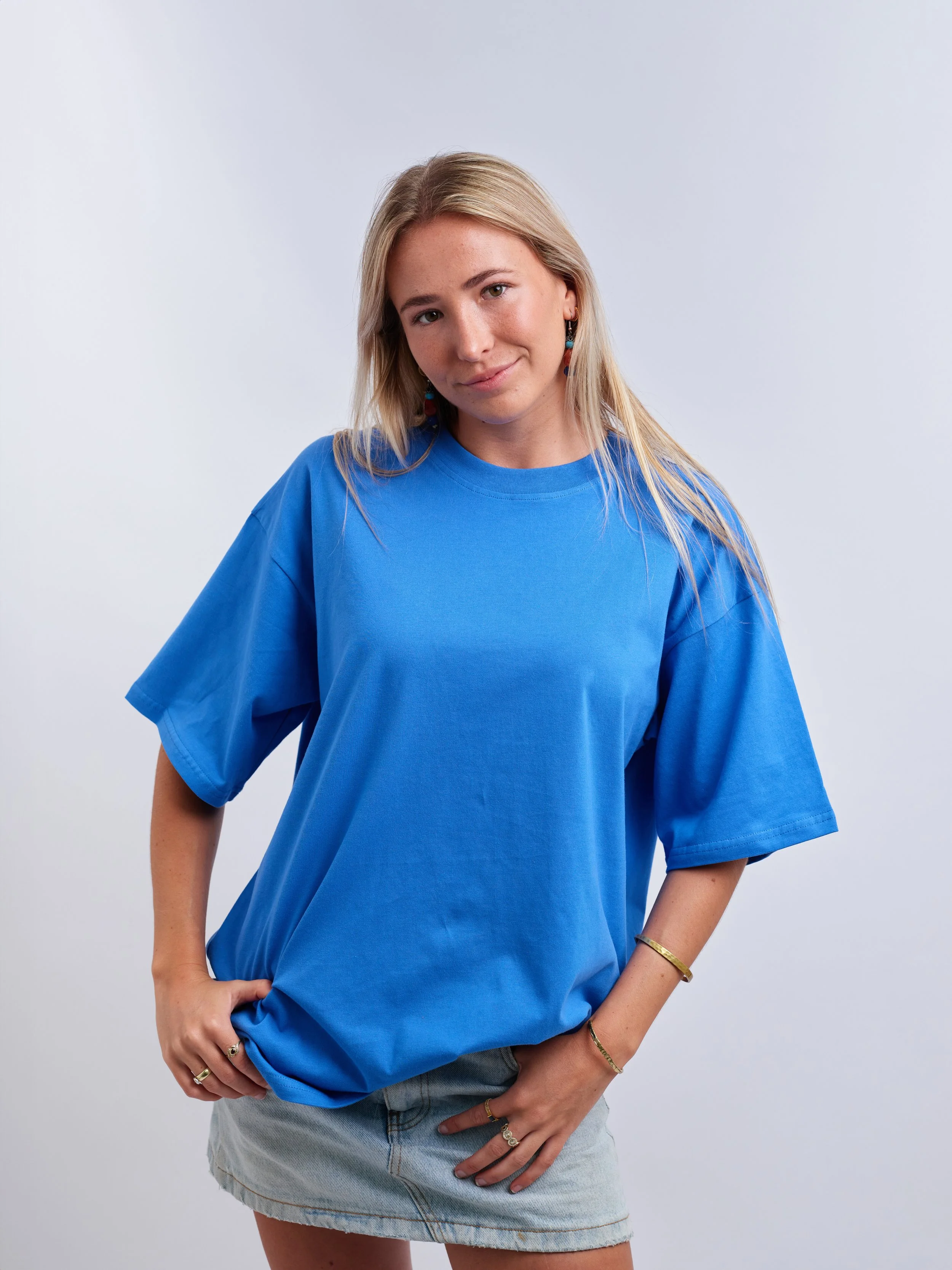 A young woman with long blonde hair wearing a loose blue t-shirt, light denim skirt, and jewelry, standing against a plain gray background. 