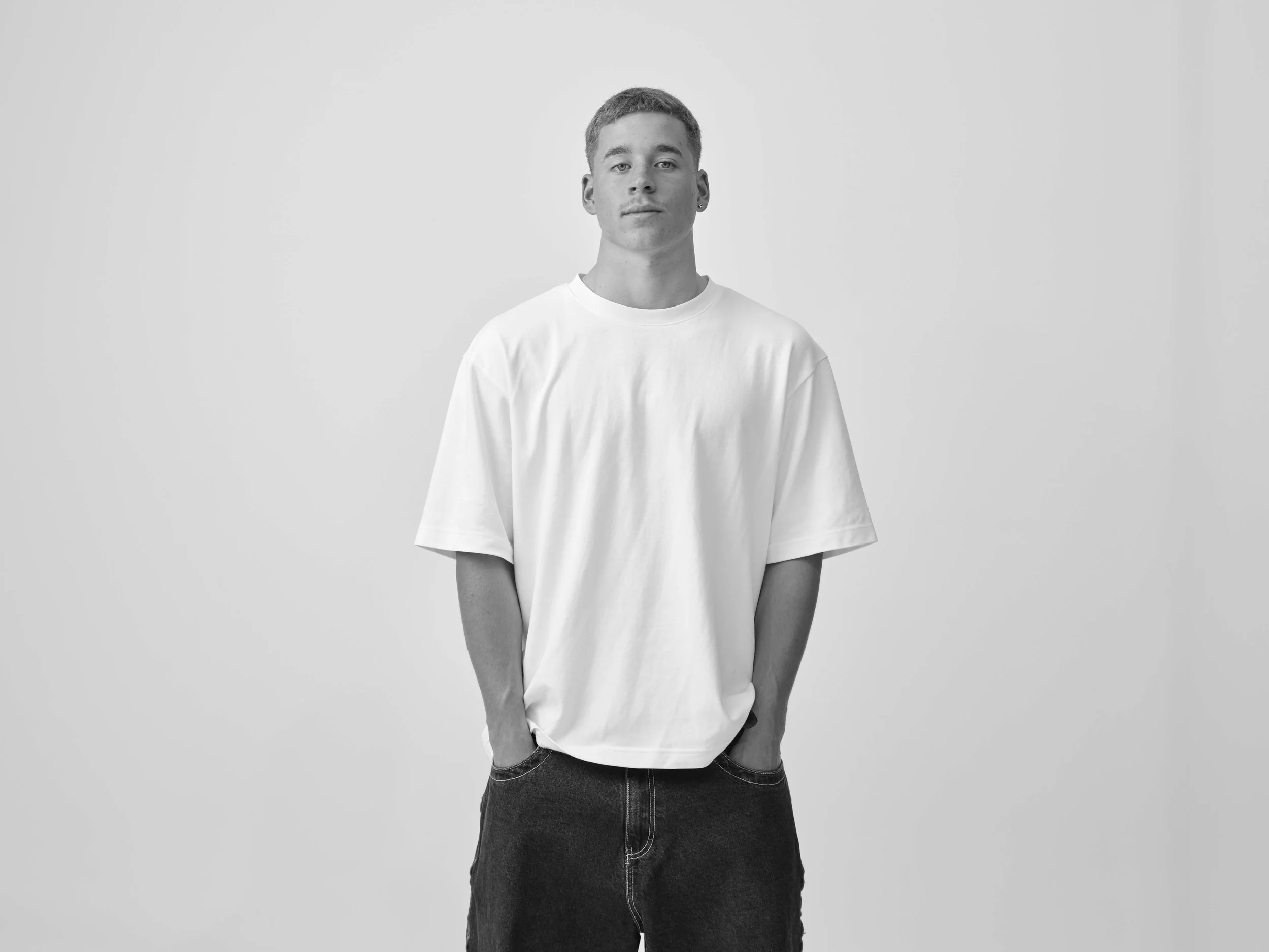 Black and white portrait of a young man standing against a plain white background, wearing a loose white t-shirt and dark pants, with hands in pockets, looking directly at the camera.