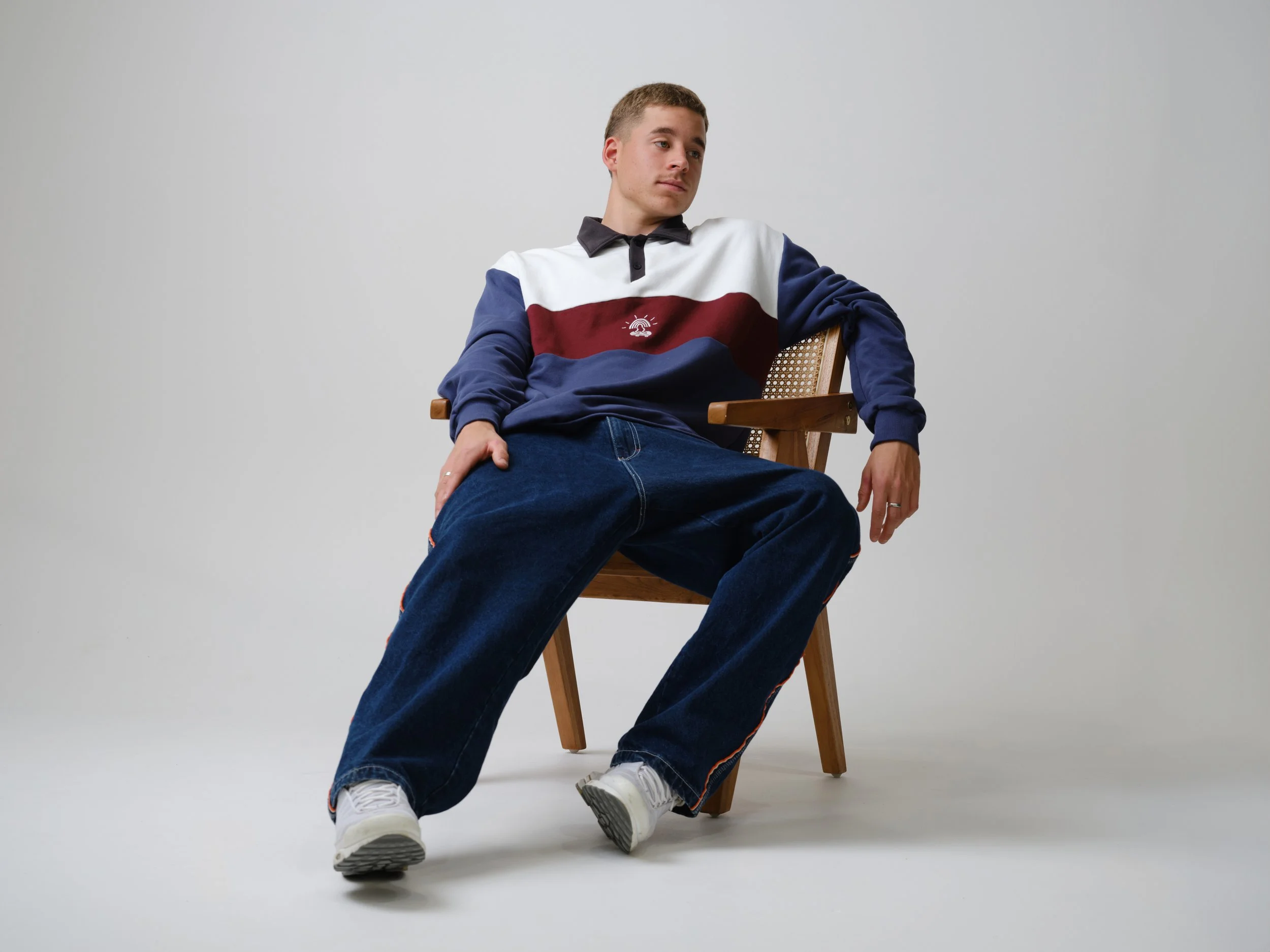 Young man seated on a wooden chair in a casual outfit, looking to the side with a relaxed expression, against a plain light background.