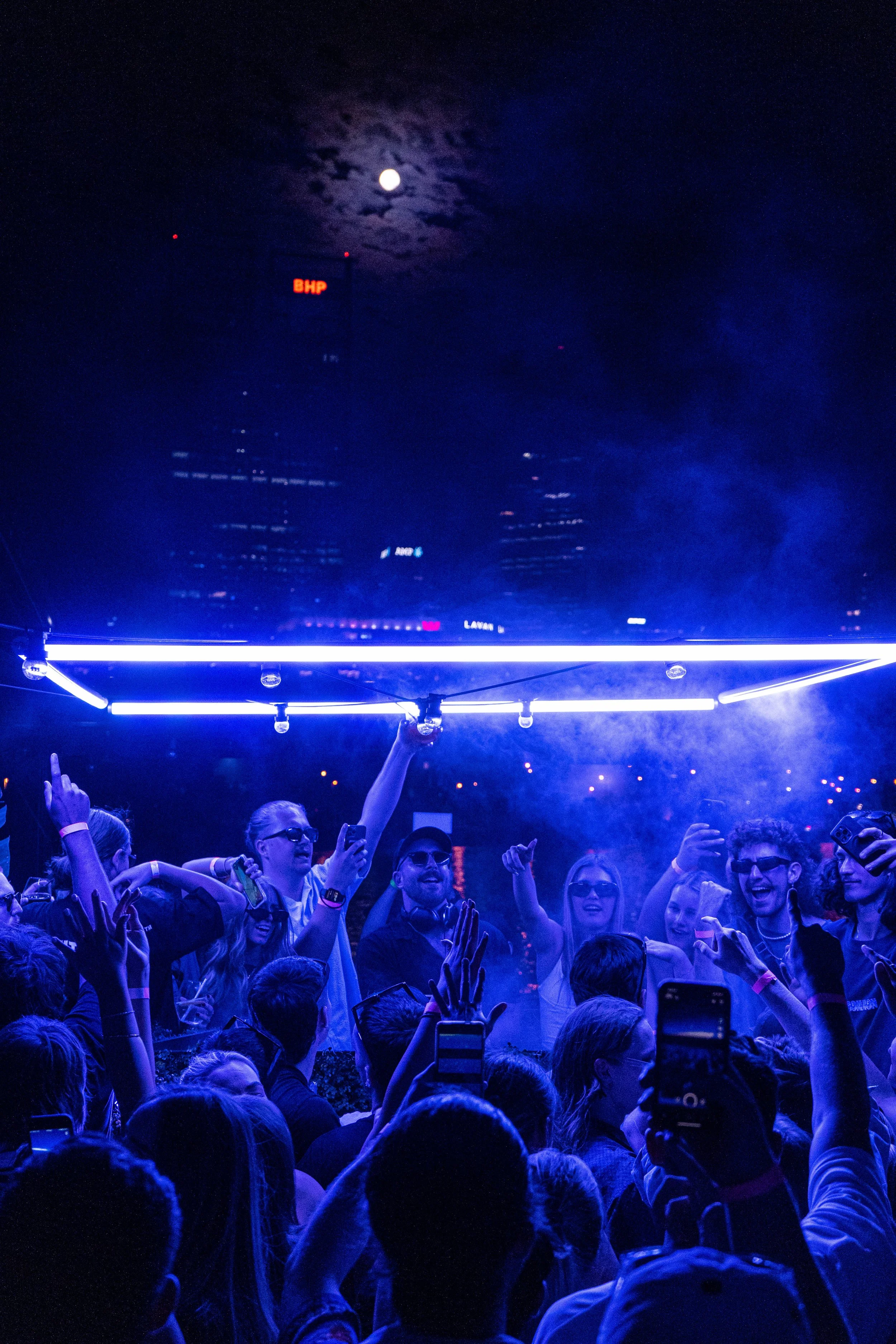 People at a nighttime outdoor concert or festival, dancing and taking photos, with a high ceiling of neon lights and a city skyline with a full moon in the cloudy sky in the background.