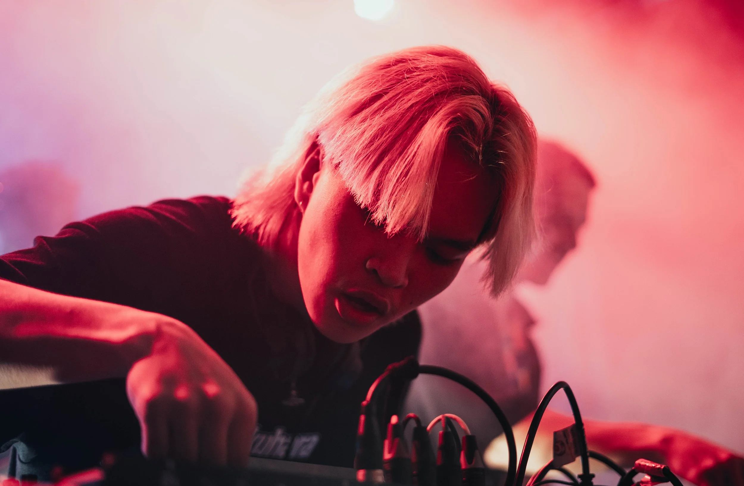 A person with bleached blonde hair is operating electronic music equipment, possibly a mixer or synthesizer, with a concentrated expression. There is a second person in the background, slightly out of focus, with pink and red lighting creating a vibr