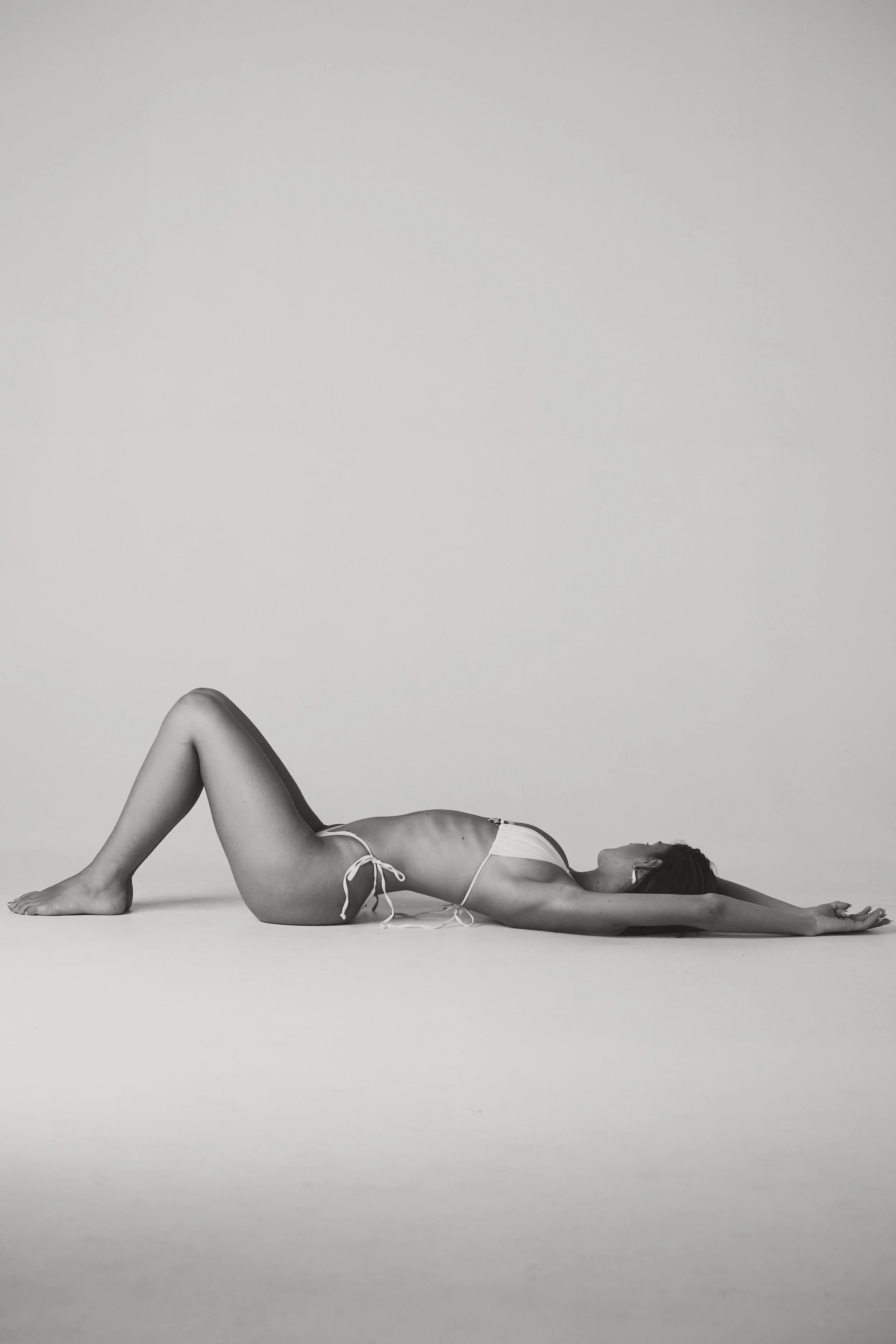 A woman lying on her back on the floor in a yoga pose, wearing a bikini, with her legs bent and arms extended overhead, on a plain background.