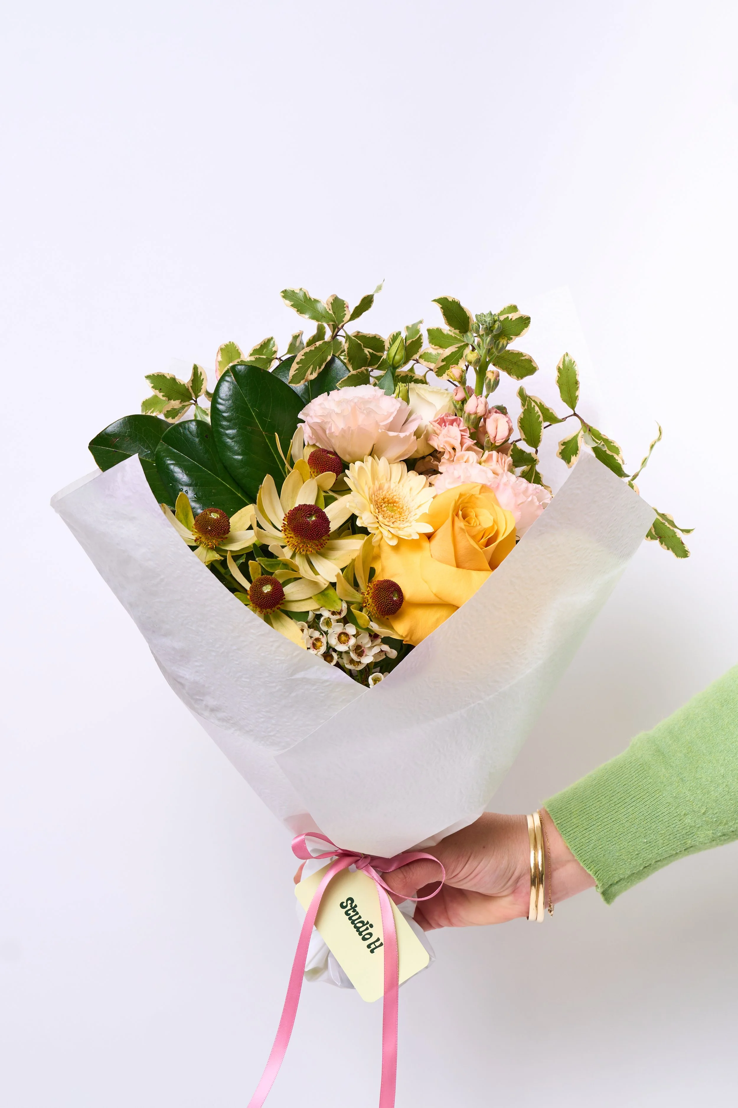 A hand holding a bouquet of mixed flowers wrapped in white paper, tied with a pink ribbon, with a yellow tag that says 'Studio H'
