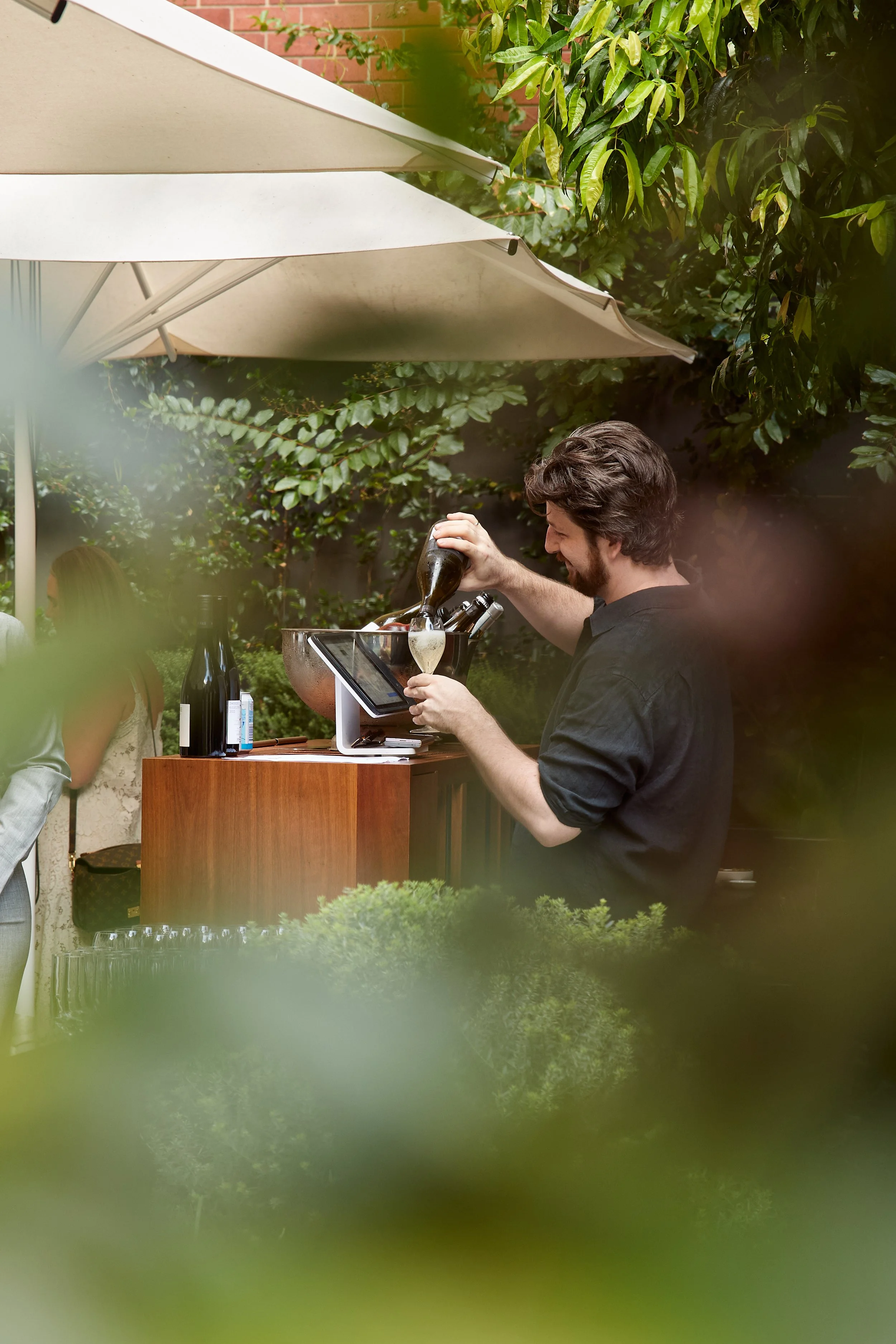 A man pouring champagne at an outdoor bar, surrounded by greenery and shade umbrellas.
