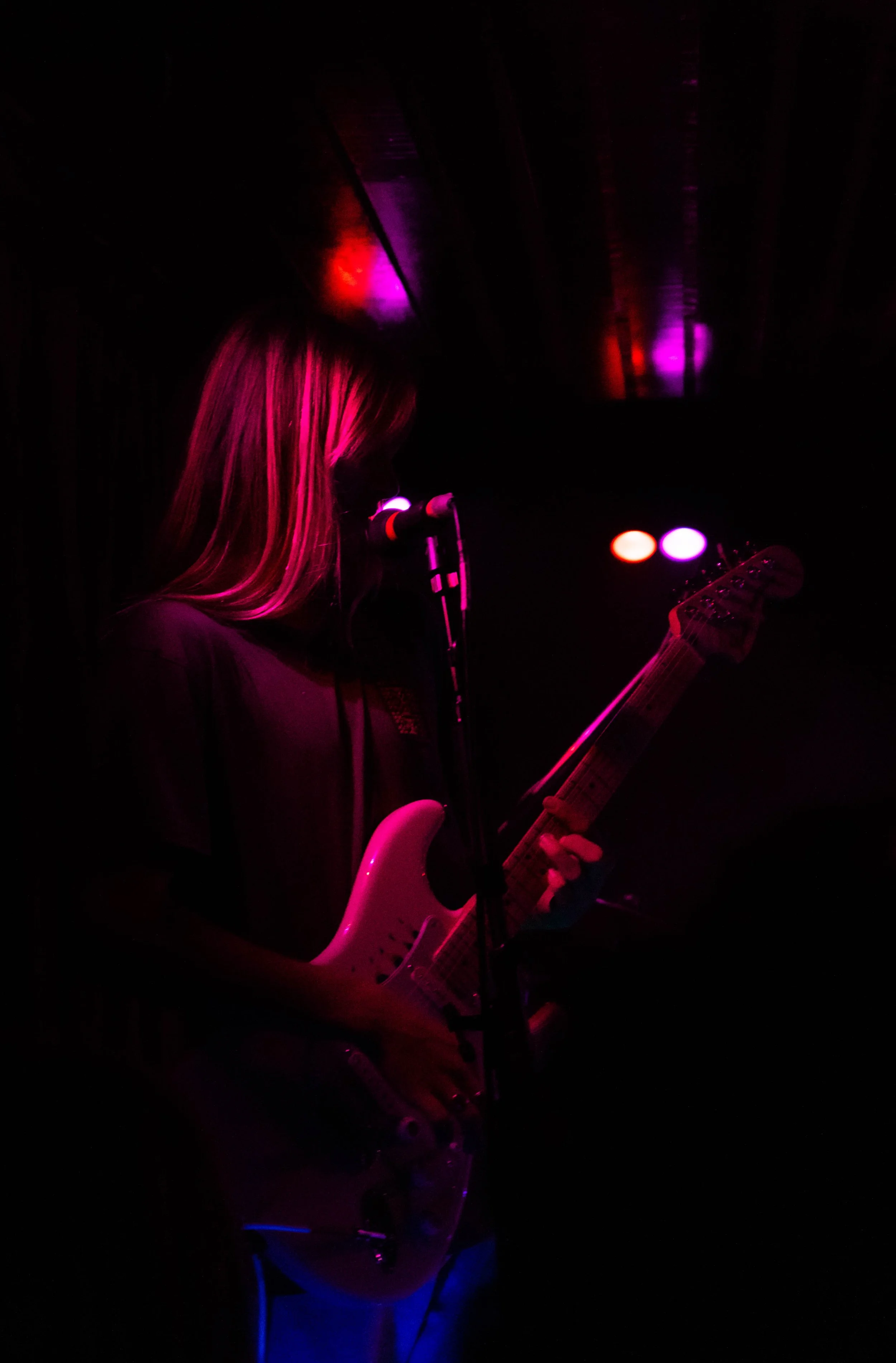 A woman playing an electric guitar on stage with dark lighting and colorful stage lights.