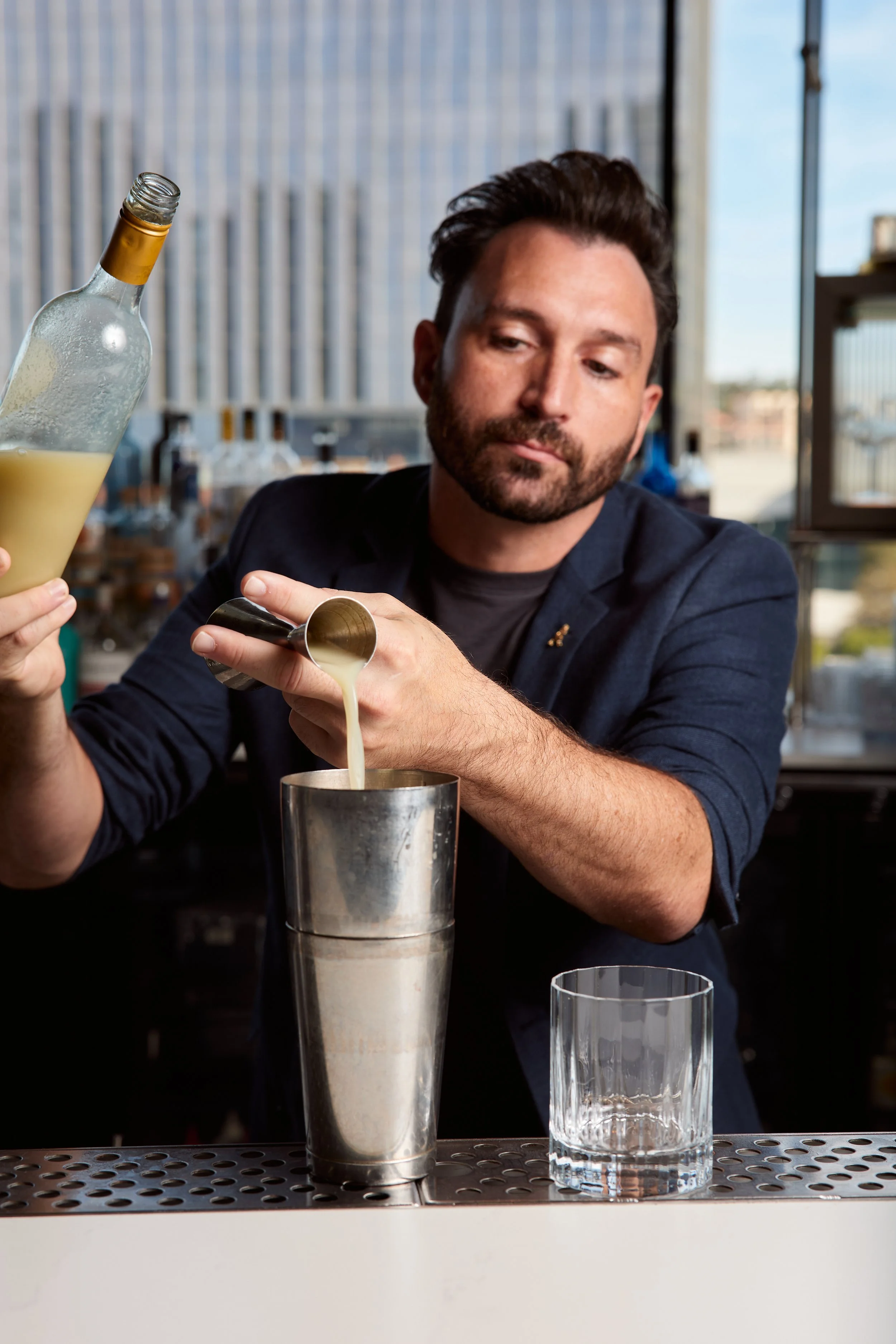 Bartender pouring a creamy cocktail from a shaker into a glass on a bar counter.