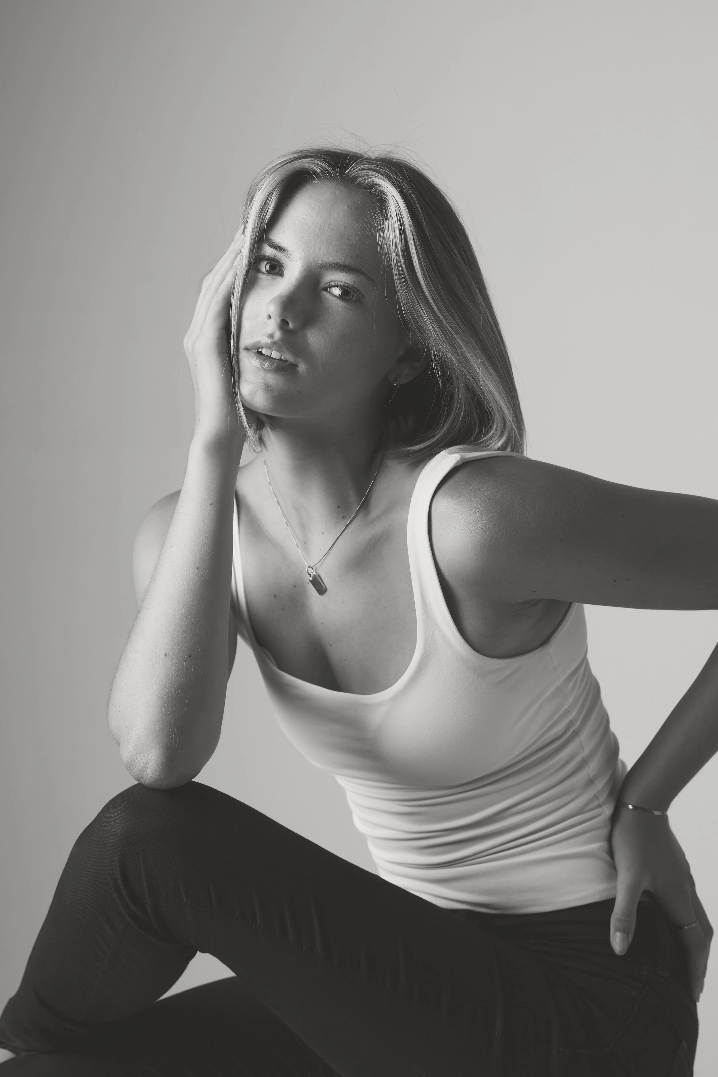 Black and white portrait of a young woman with straight hair, wearing a white tank top and dark pants, seated with one hand on her head and looking at the camera.