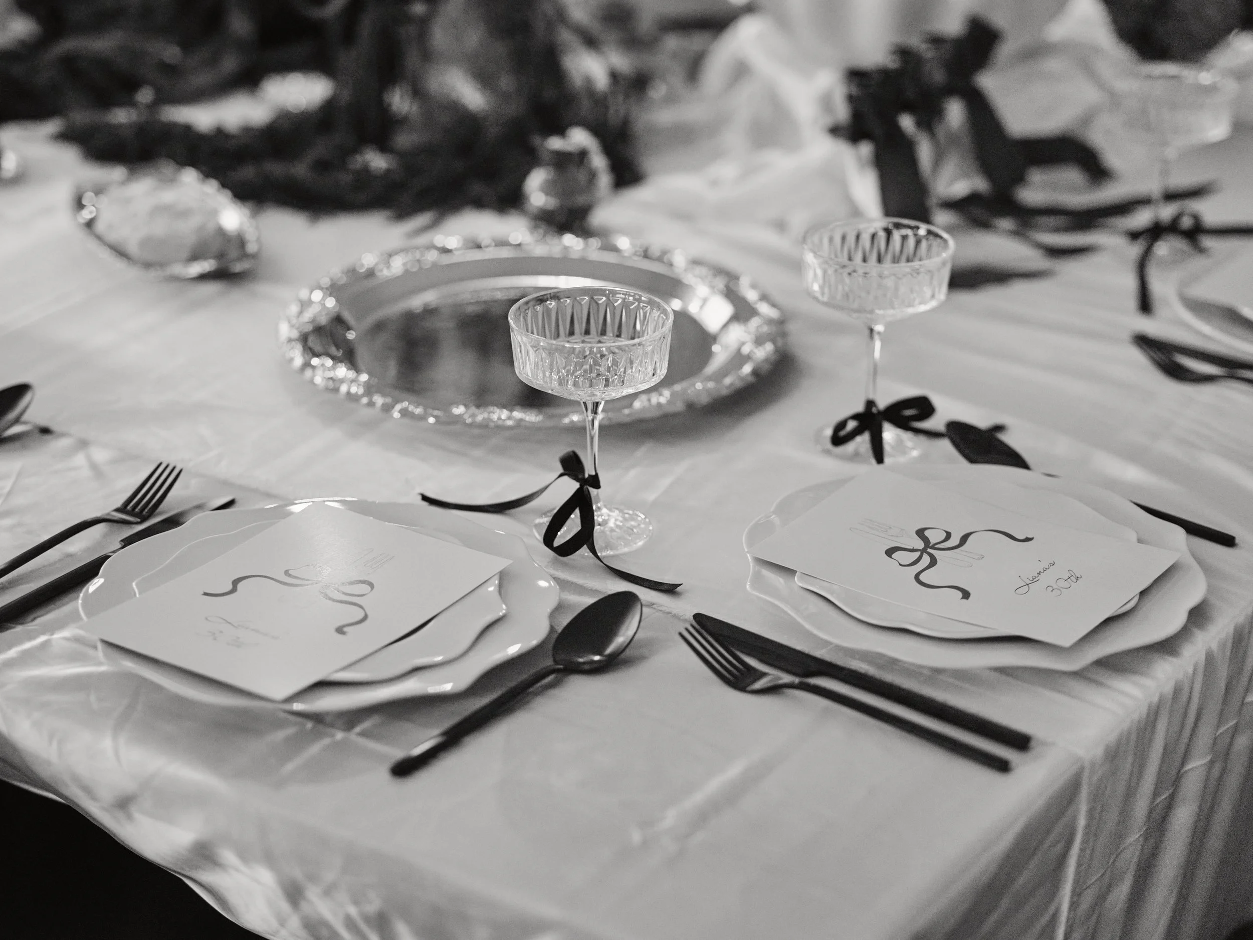 A formal dinner table setting with white plates, silverware, two decorative drink glasses with black ribbons, plates with handwritten names, and a silver centerpiece plate on a white tablecloth.