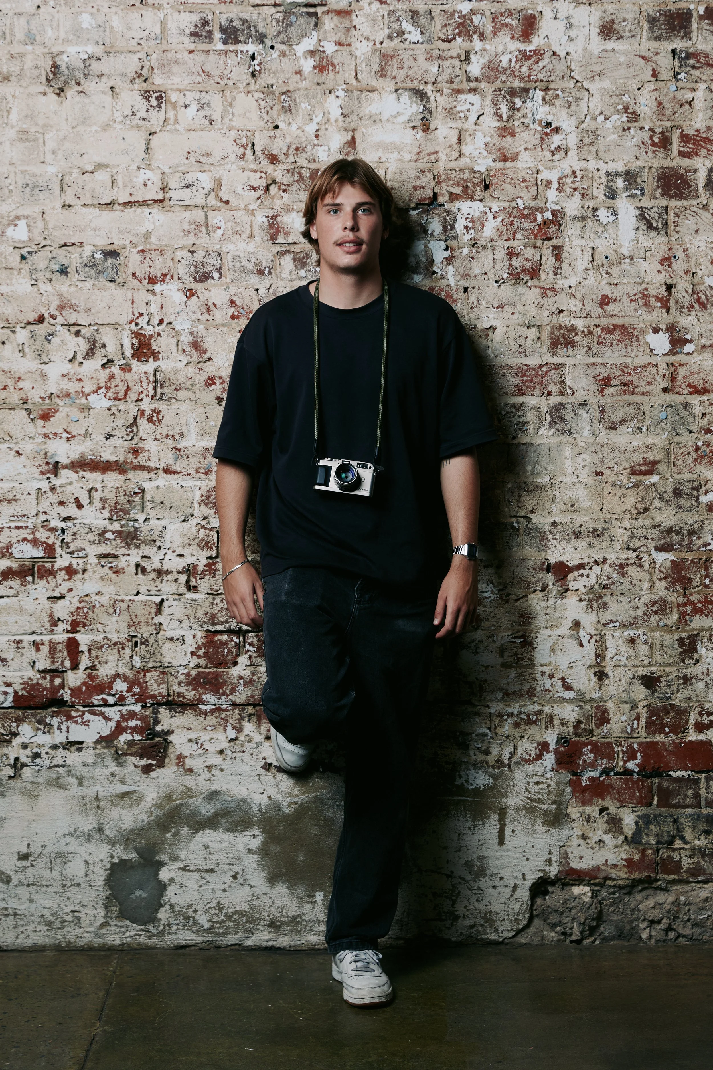 A young man with light skin and brown hair standing against a weathered brick wall. He is dressed in a black T-shirt, black pants, and white sneakers, with a camera hanging around his neck. He has a watch on his left wrist and a bracelet on his right wrist.