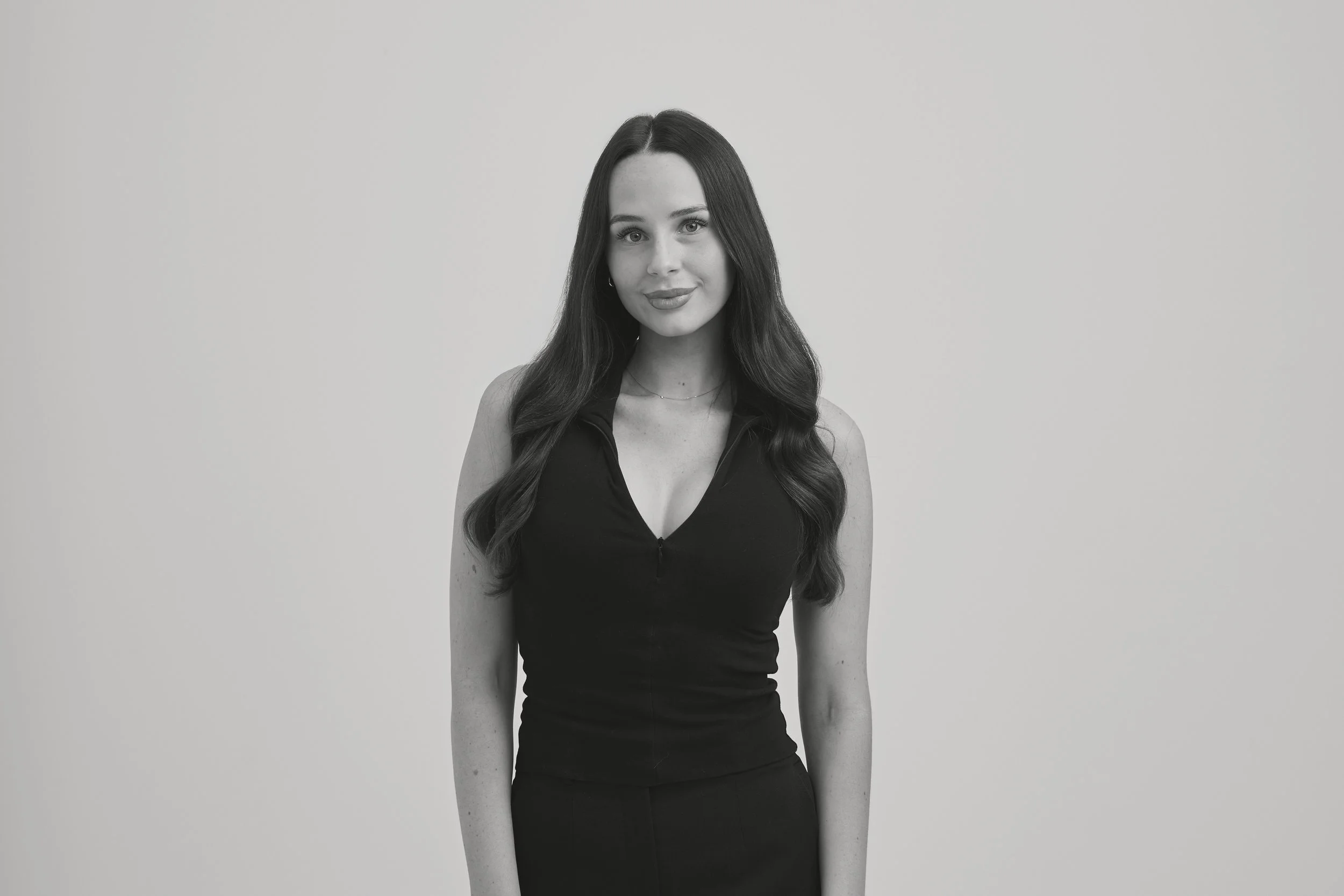 Black and white portrait of a woman with long wavy hair, wearing a sleeveless zip-up top, smiling lightly, standing against a neutral background.