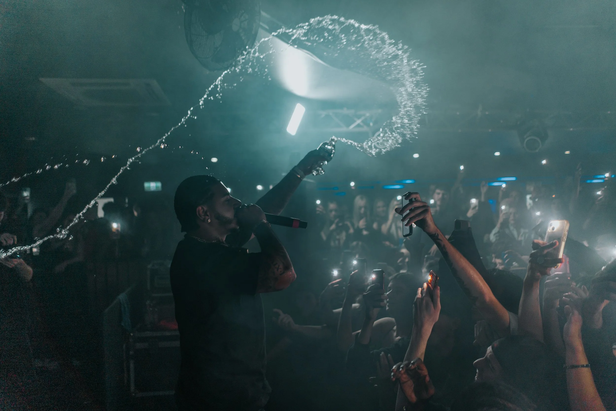 Performer holding a microphone in a dark club, spraying water into the air as crowd members record the moment with their phones.