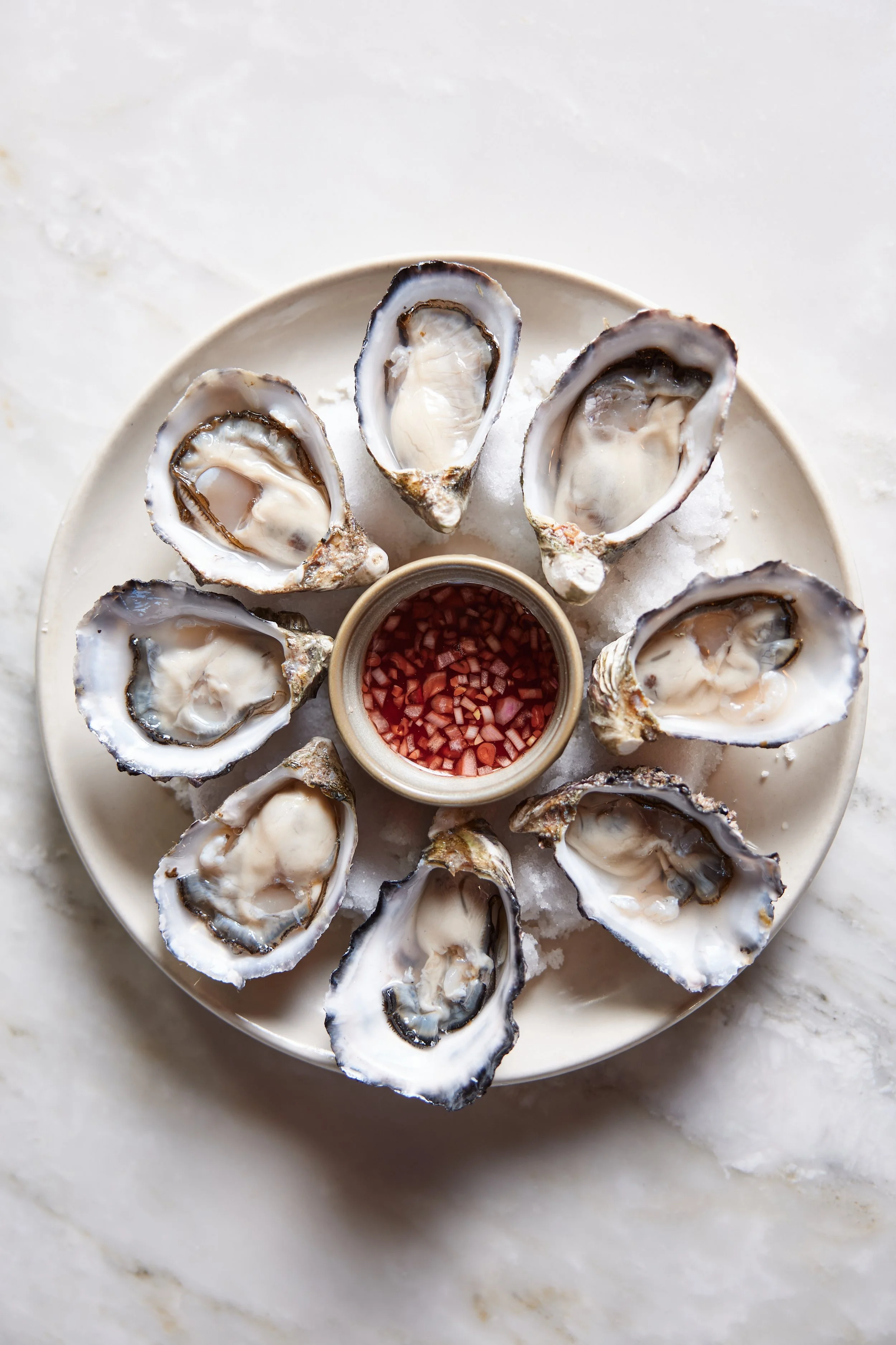 A white plate with eight open oysters on the half shell arranged in a circle on ice, with a small bowl of red cocktail sauce in the center.