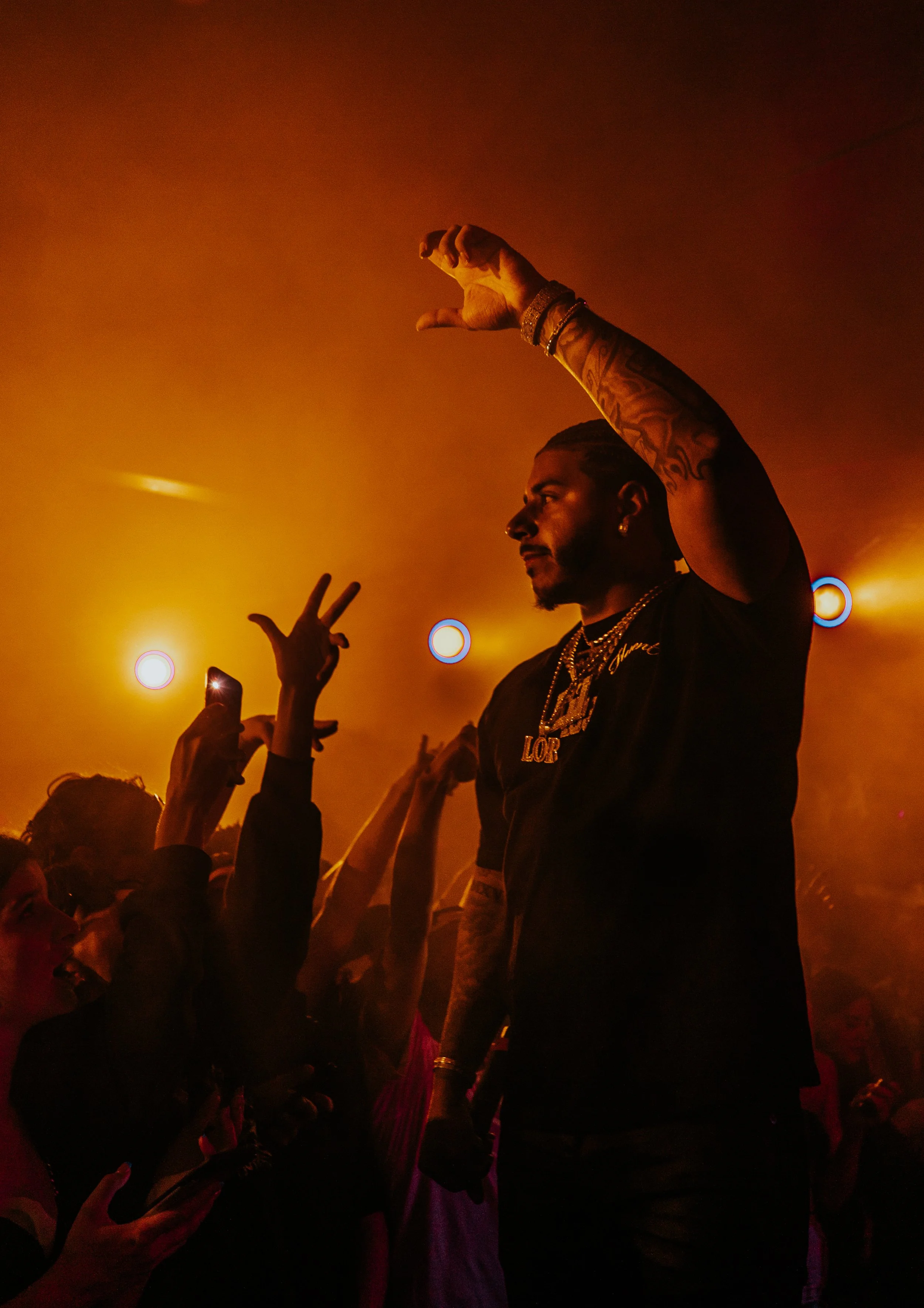 A male performer with tattoos, wearing chains and jewelry, performing on stage with orange lighting, surrounded by fans taking photos and reaching out.