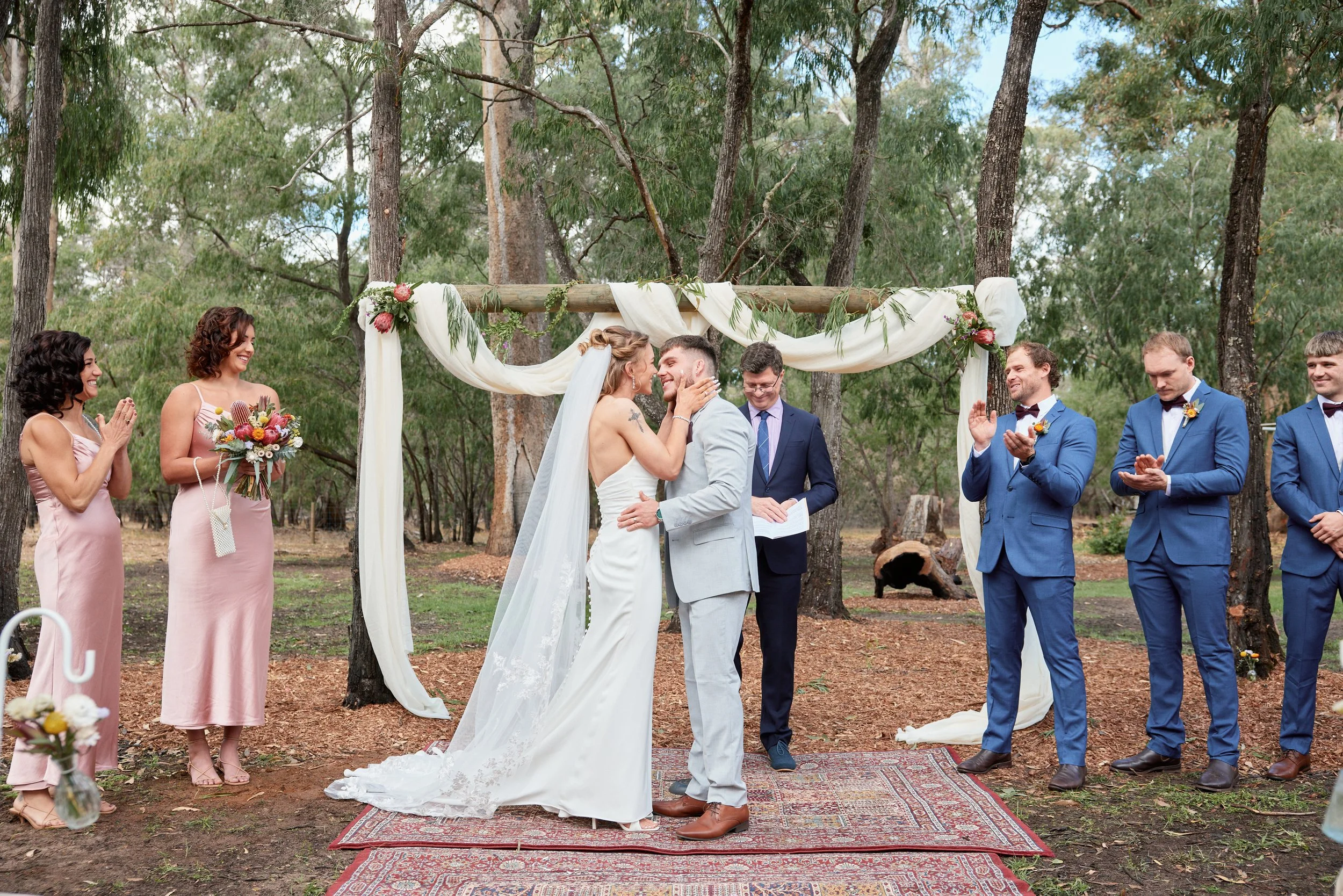 A bride and groom in wedding attire share a kiss under an outdoor wedding arch decorated with white fabric and flowers, with bridesmaids and groomsmen clapping and smiling in a wooded area.