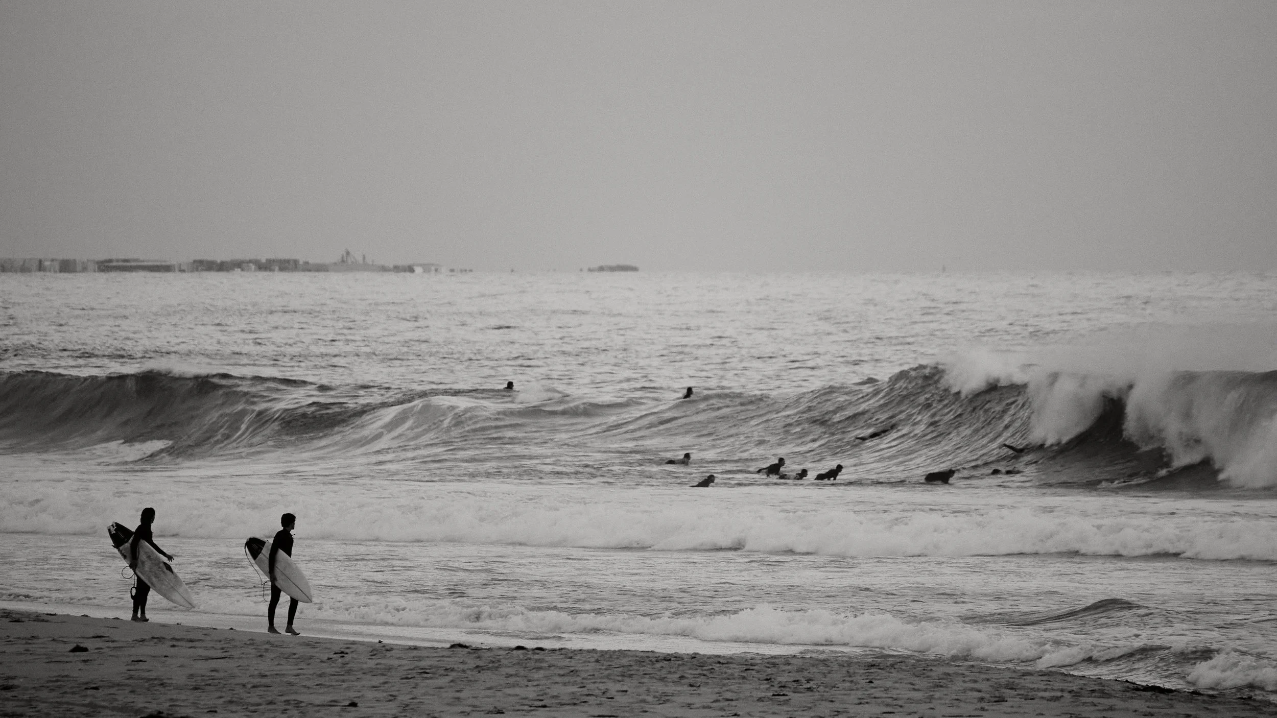 Two surfers walking on the beach carrying surfboards, with surfers in the water riding waves, in black and white.
