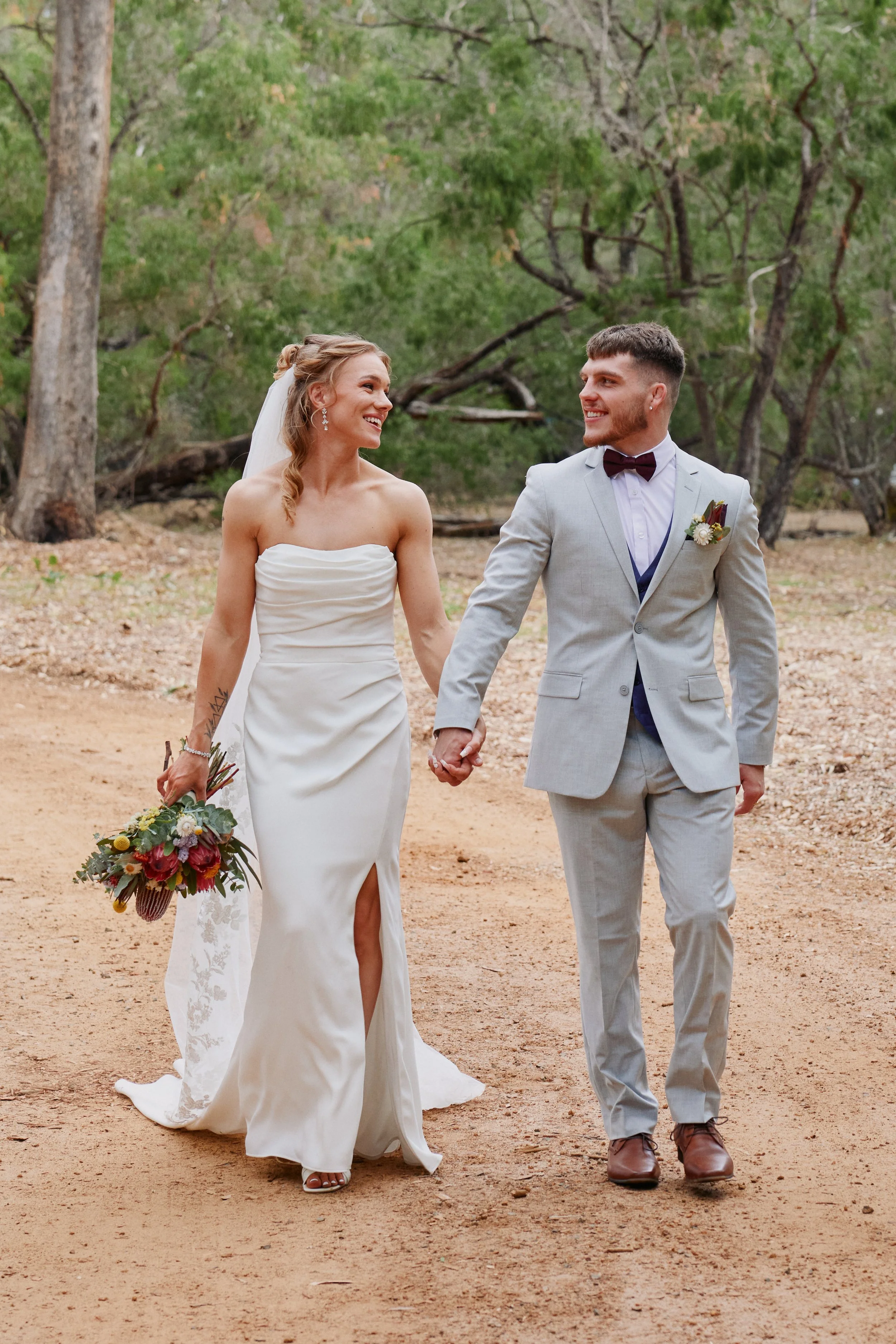 A bride and groom walking hand in hand outdoors, smiling at each other, with trees and natural surroundings in the background.