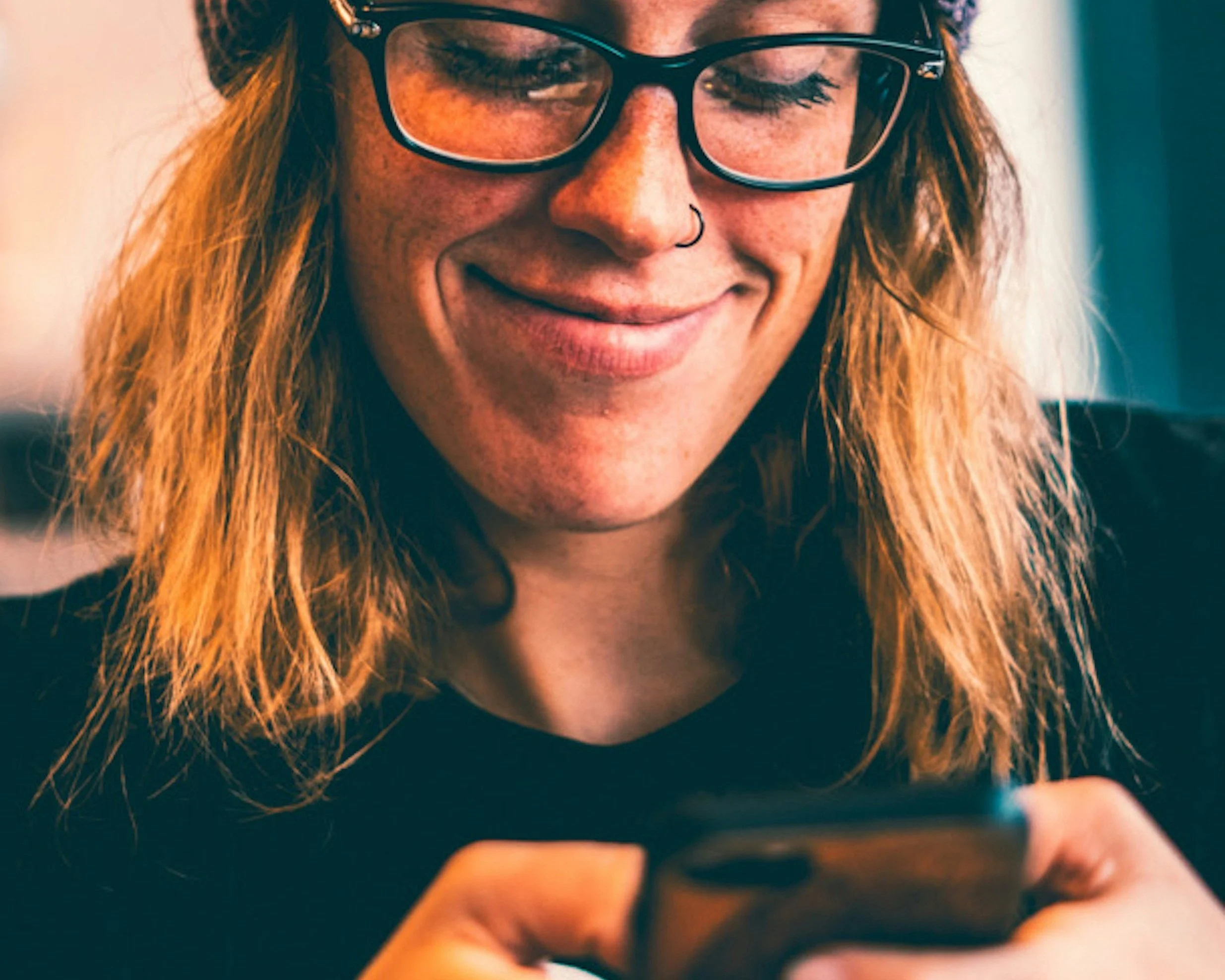 A woman with red hair, glasses, and a nose ring smiling while looking at her phone.