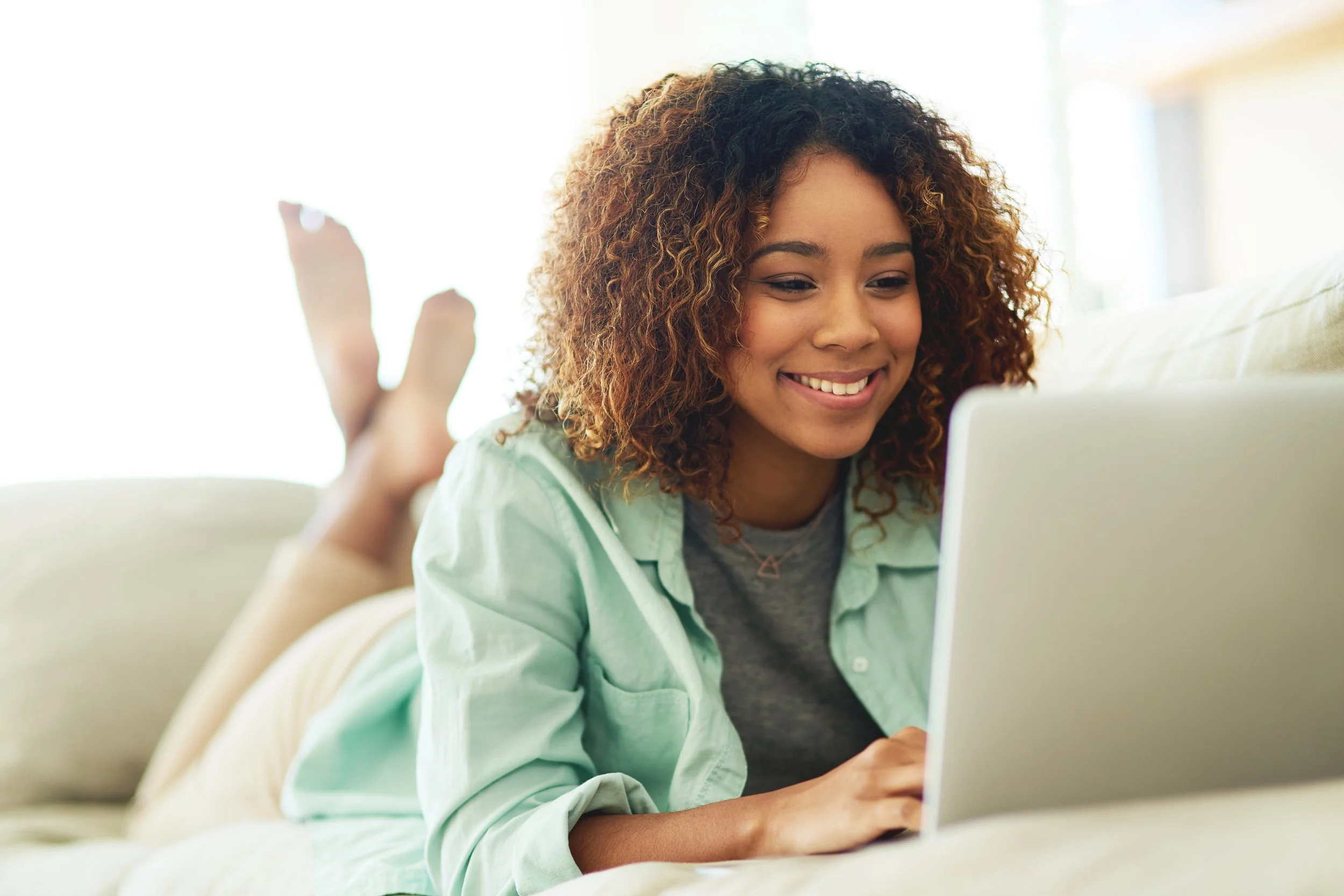 A young woman with curly hair smiling while lying on her stomach on a beige sofa, using a laptop in a bright room.