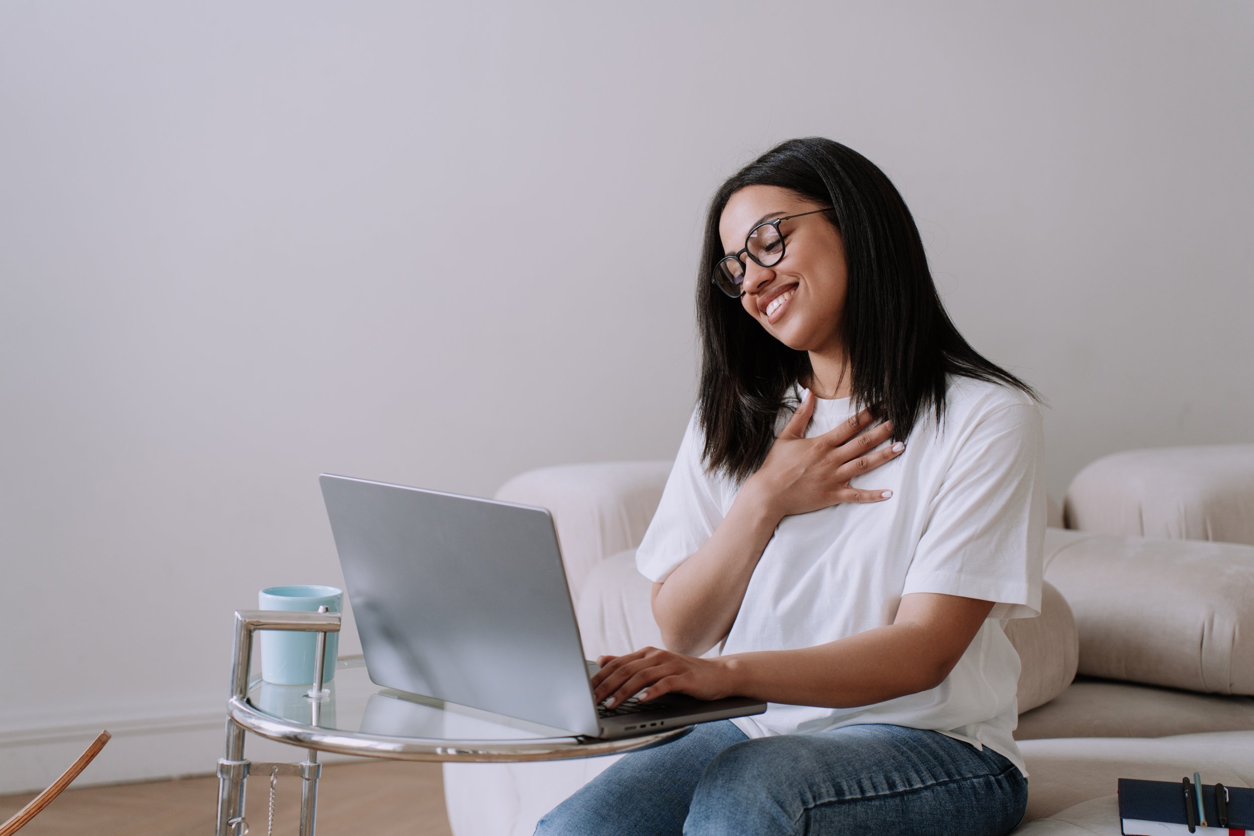 A woman with glasses and long dark hair, sitting on a white sofa, smiling and touching her chest, while using a silver laptop. A small round side table with a mug and a notepad with a pen are nearby.