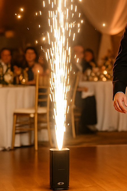 A firework sparkler shooting sparks into the air at a celebration or wedding reception, with guests seated at decorated tables in the background.