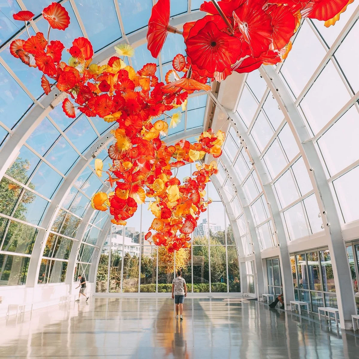 A glass atrium with an art installation of hanging red, orange, and yellow glass flowers or leaves, with a person walking underneath and sunlight streaming through.