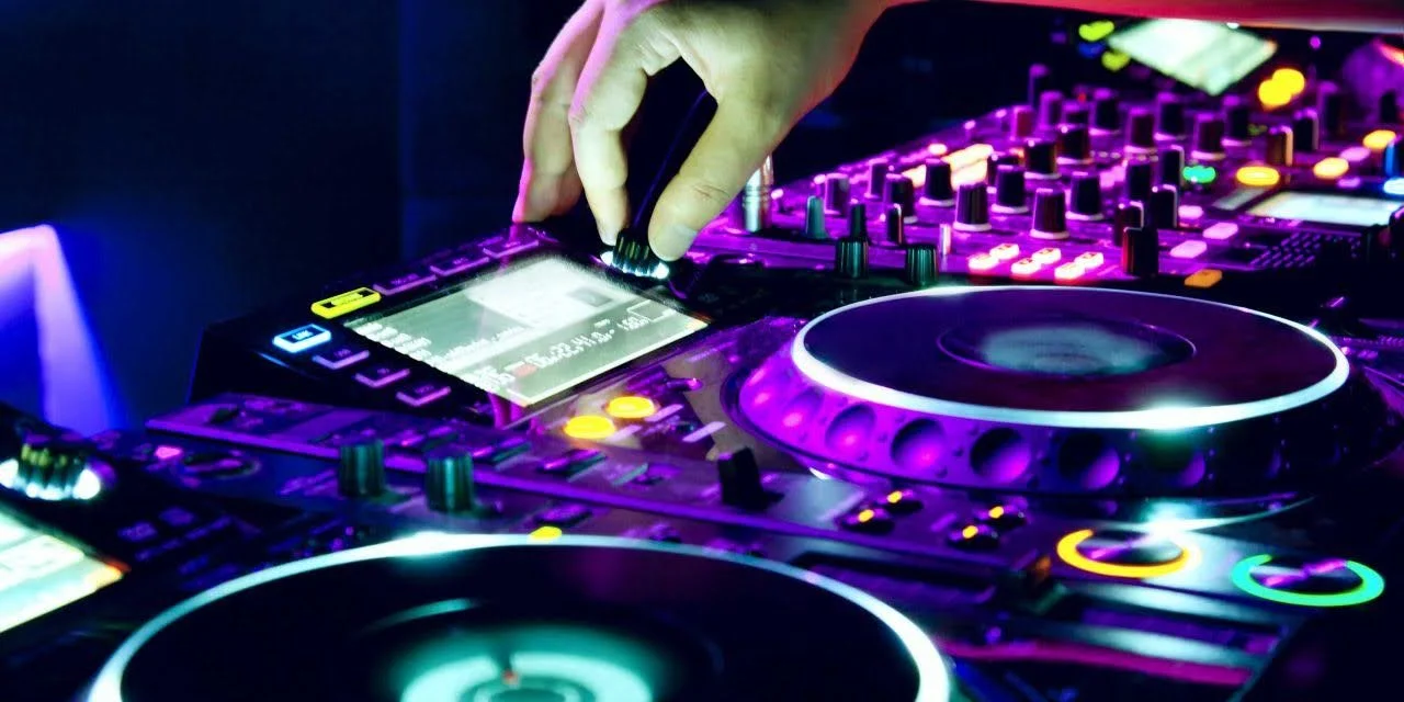 Close-up of a DJ mixing music on a digital DJ controller with colorful backlit buttons and knobs, illuminated in neon purple, blue, and yellow lights.