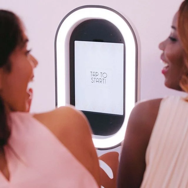 Two women smiling while facing a mirror with a touchscreen display that reads 'TAP TO START!'