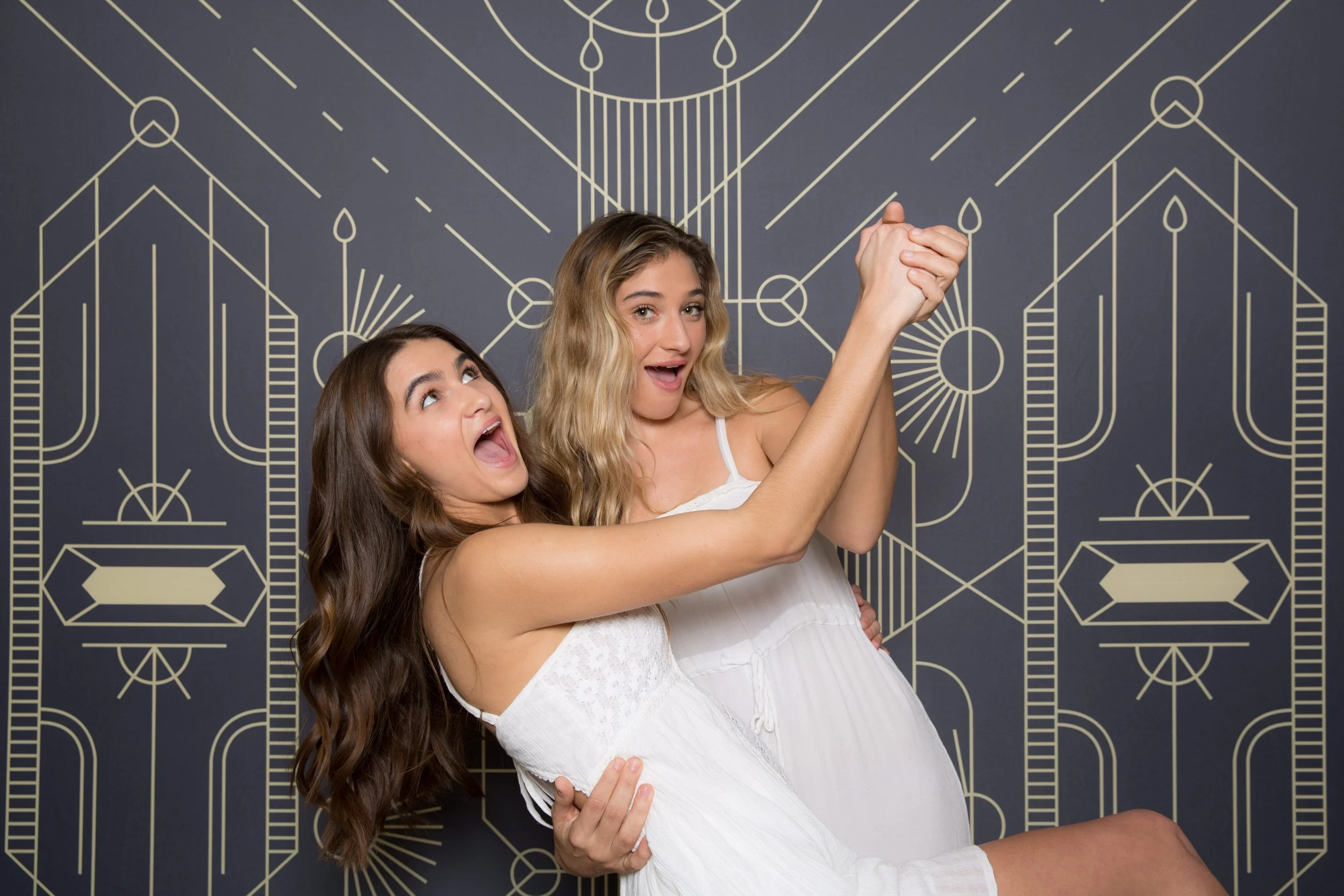 Two young women in white dresses celebrating, one lifting the other in front of a gray background with yellow geometric designs.