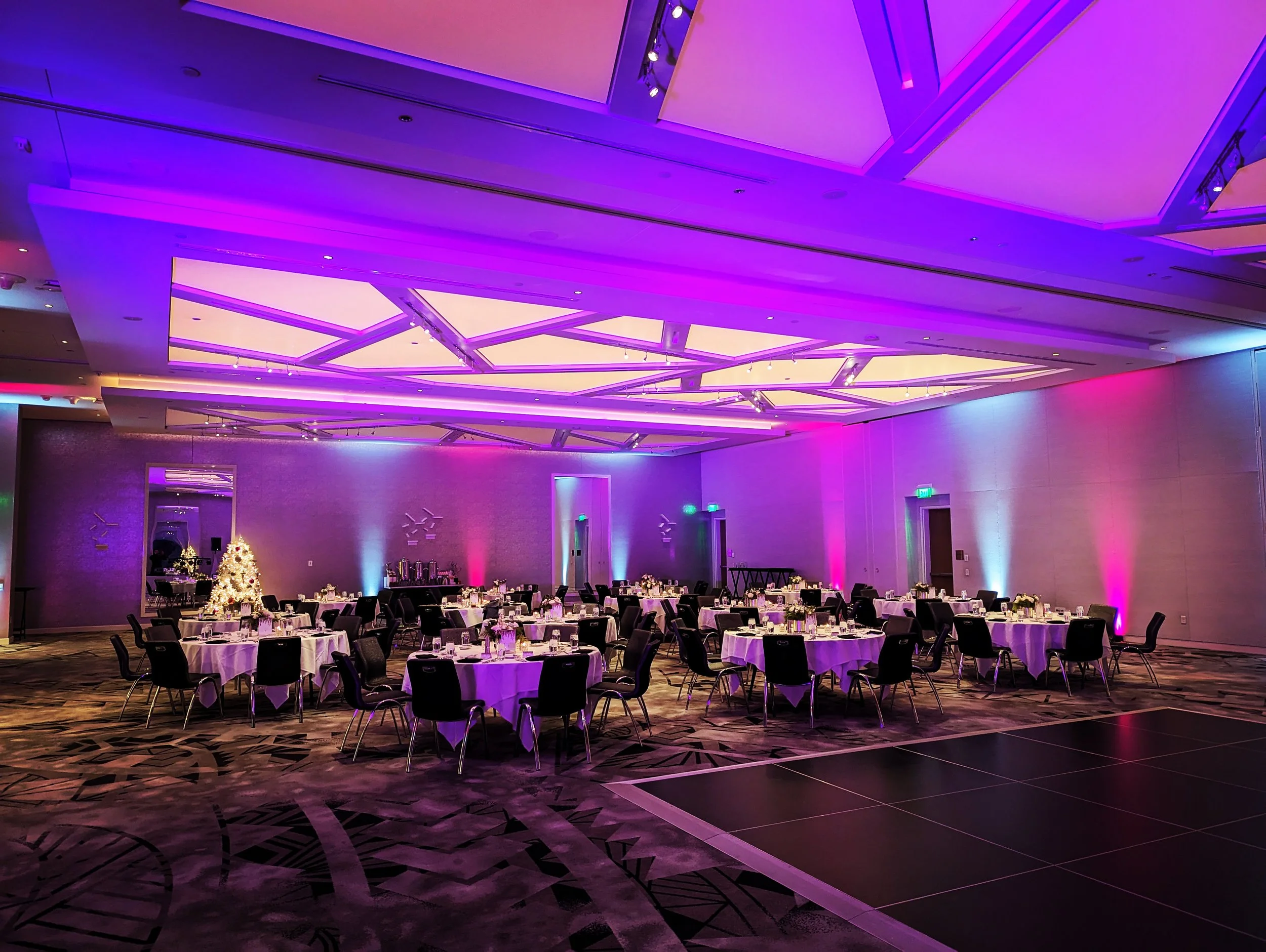 An elegant event hall decorated with purple and pink lighting, tables with white tablecloths and floral centerpieces, and a Christmas tree in the corner.
