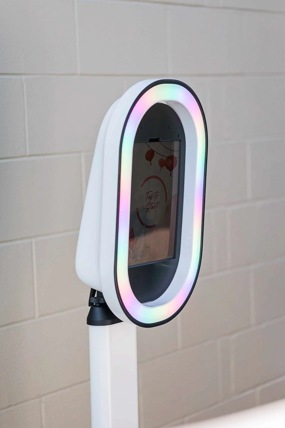 A white circular device with a rainbow-colored LED ring around a screen displaying the words "Tap to Start". The device is mounted on a stand against a beige brick wall.