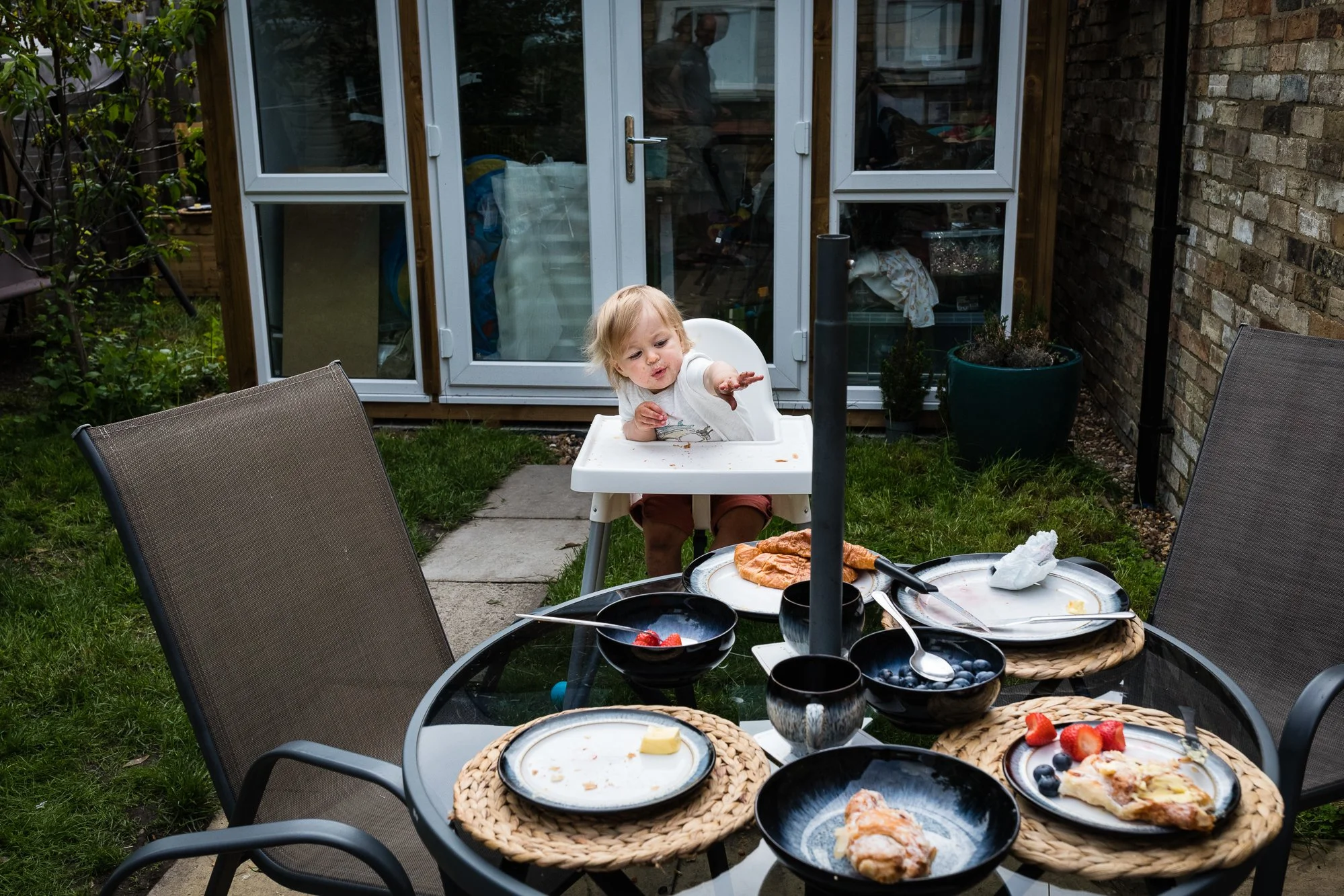 Child sitting in a high chair reaching out with food on the tray in a backyard dining area with a glass table, various plates of partially eaten food and bowls of berries, surrounded by outdoor chairs, with a garden and a brick wall in the background