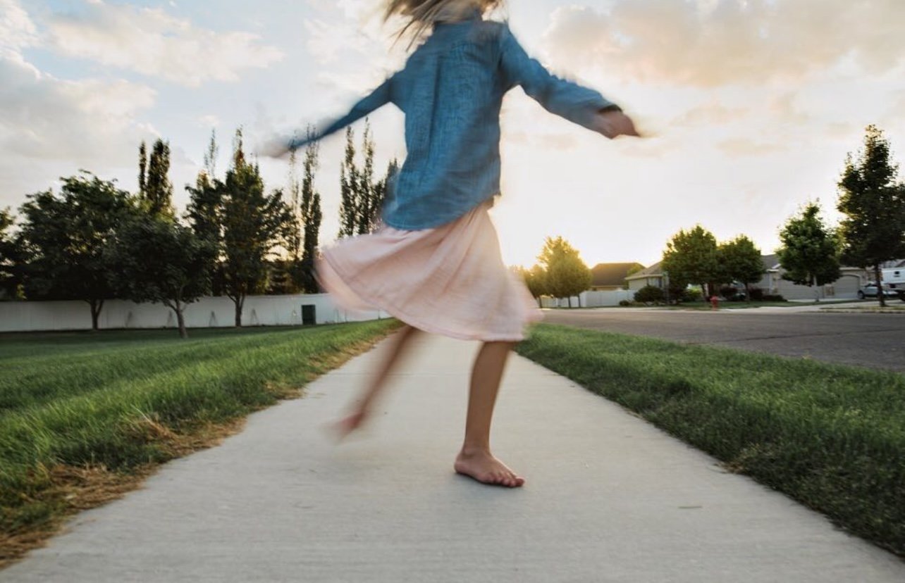 A girl in a pink skirt and blue jacket spinning on a sidewalk at sunset with trees and houses in the background.