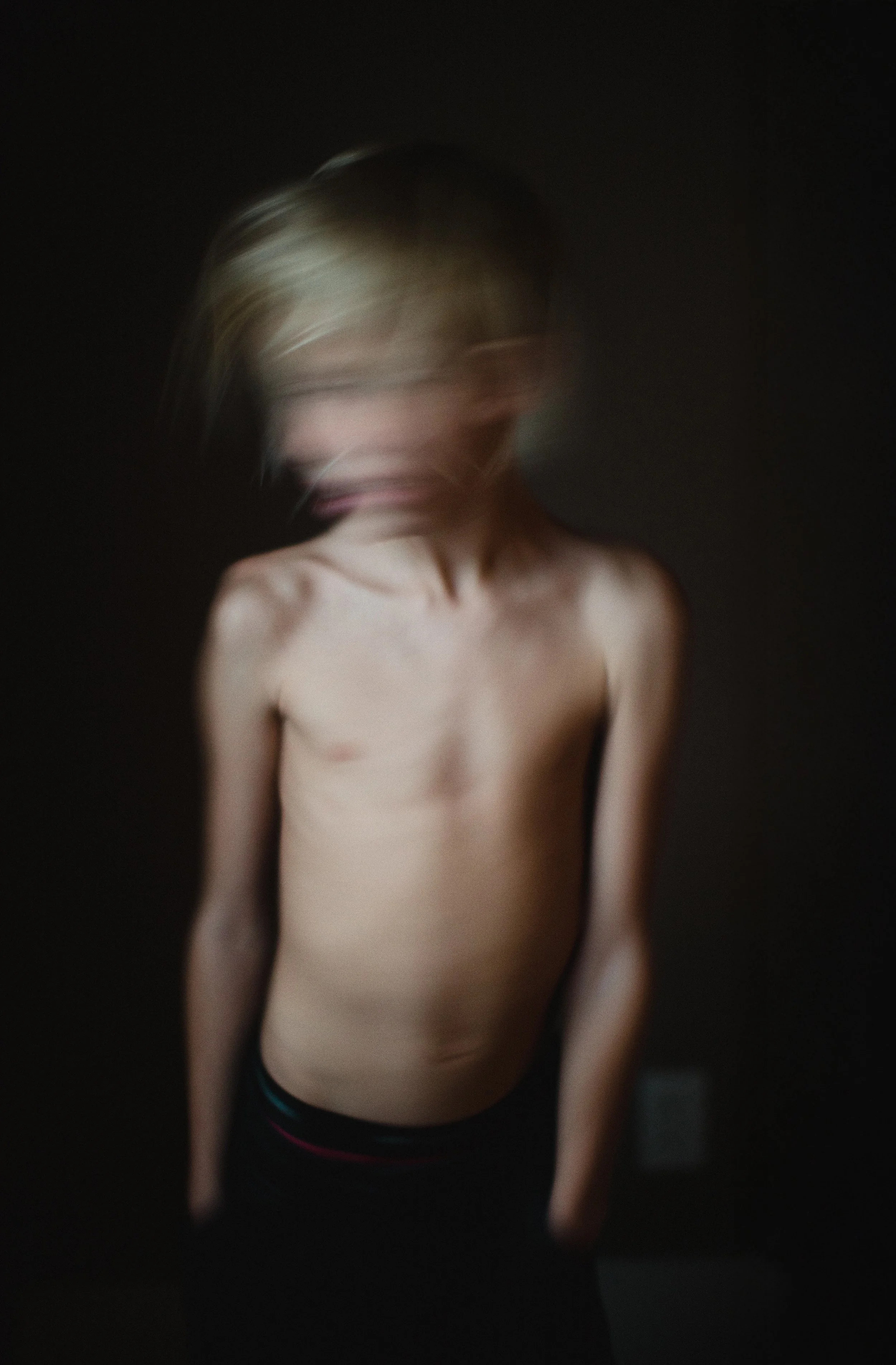 A young boy with blonde hair, shirtless, standing against a dark background, with a motion-blurred face and head.