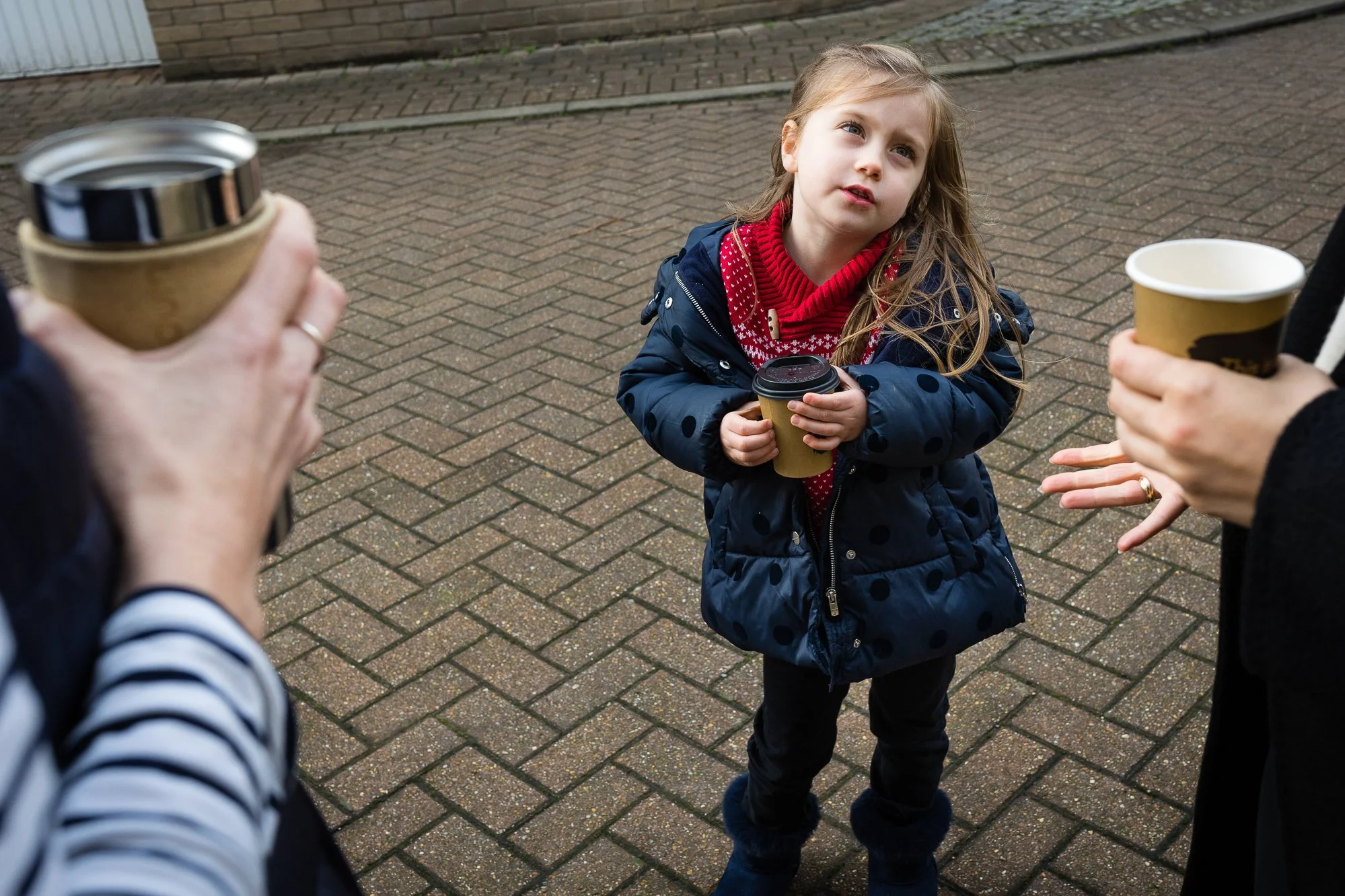 A young girl with blonde hair holding a paper coffee cup, standing on a paved brick sidewalk, surrounded by two adults also holding coffee cups, with a wooden fence and a building in the background.
