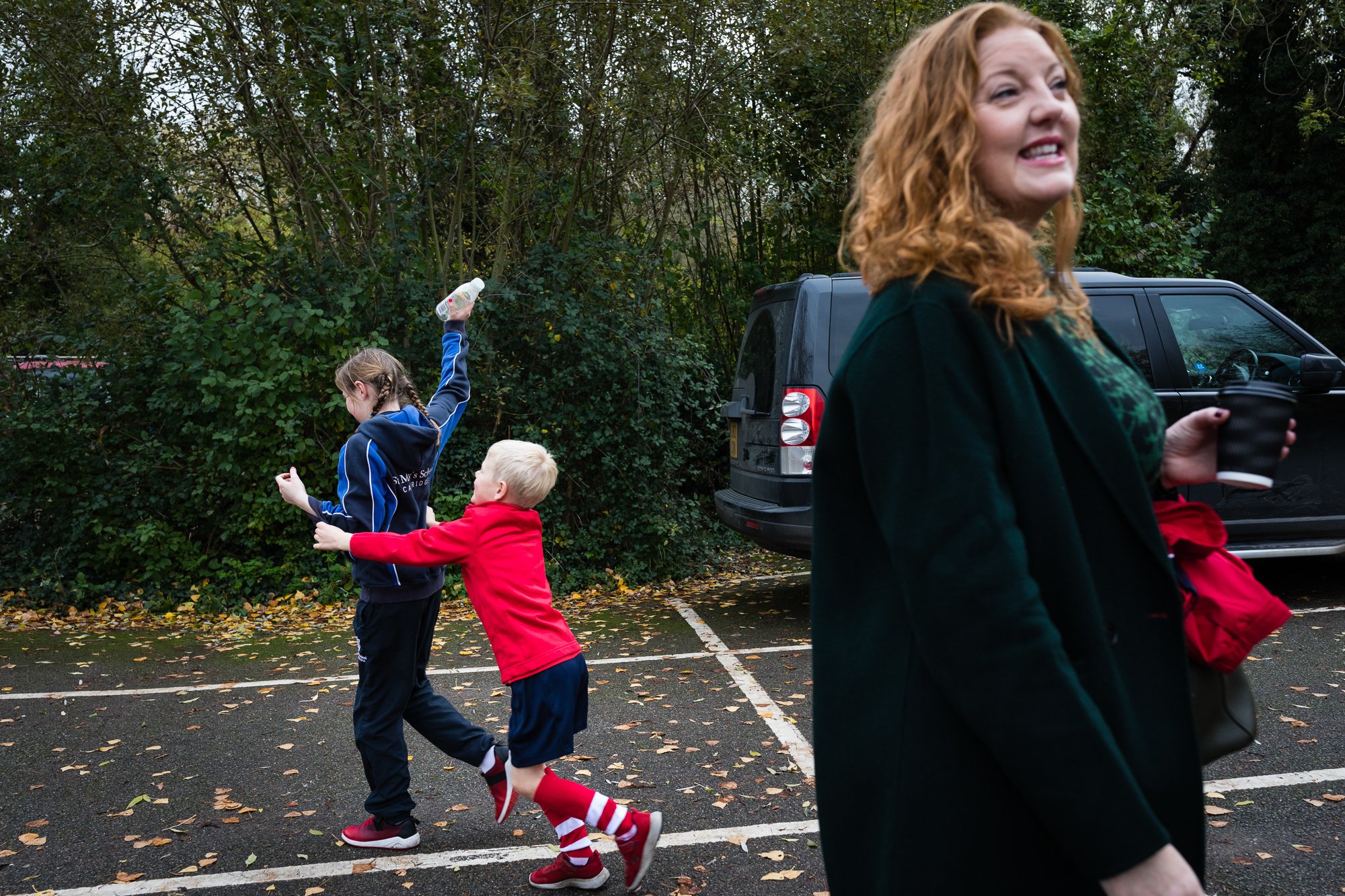 Two young children, a girl and a boy, playing in a parking lot with a woman watching nearby. The girl is tossing an object into the air while the boy tries to catch it. The woman is holding a coffee cup and smiling.