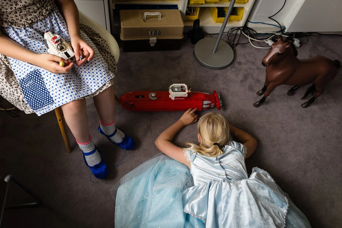 Two children playing with LEGO boats on the carpeted floor of a room, one sitting on a chair and the other lying down in a light blue dress, with a plastic horse toy nearby.