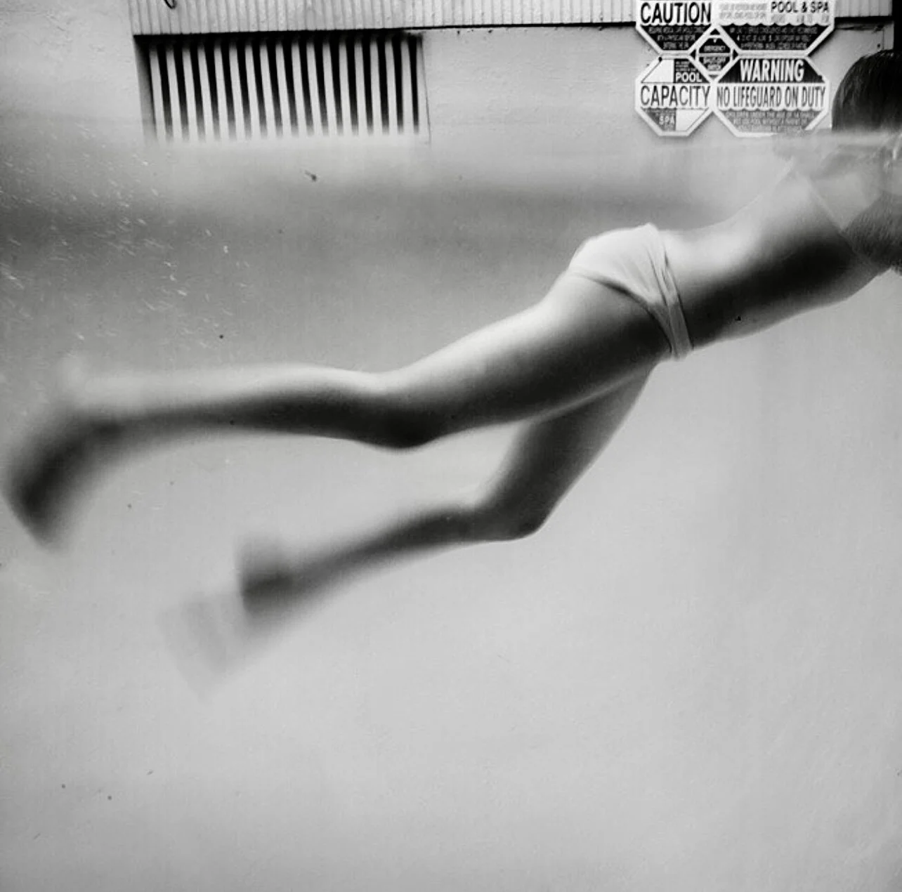 A person swimming underwater in a pool, wearing a bathing suit. Signs and a striped vent are visible on the wall in the background.