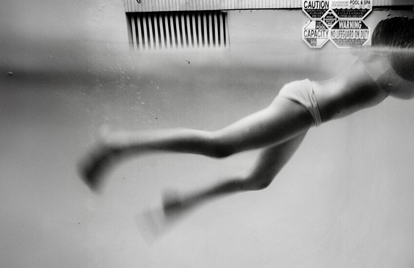 A person with light skin swimming underwater, wearing a tank top and shorts, with safety and pool warning signs on the wall in the background.