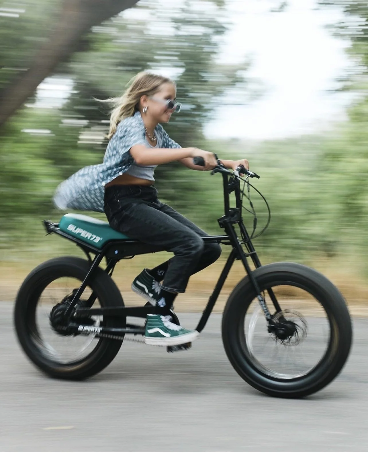 Young woman riding an electric mountain bike on a paved path, mid-motion, outdoors with green trees in background.