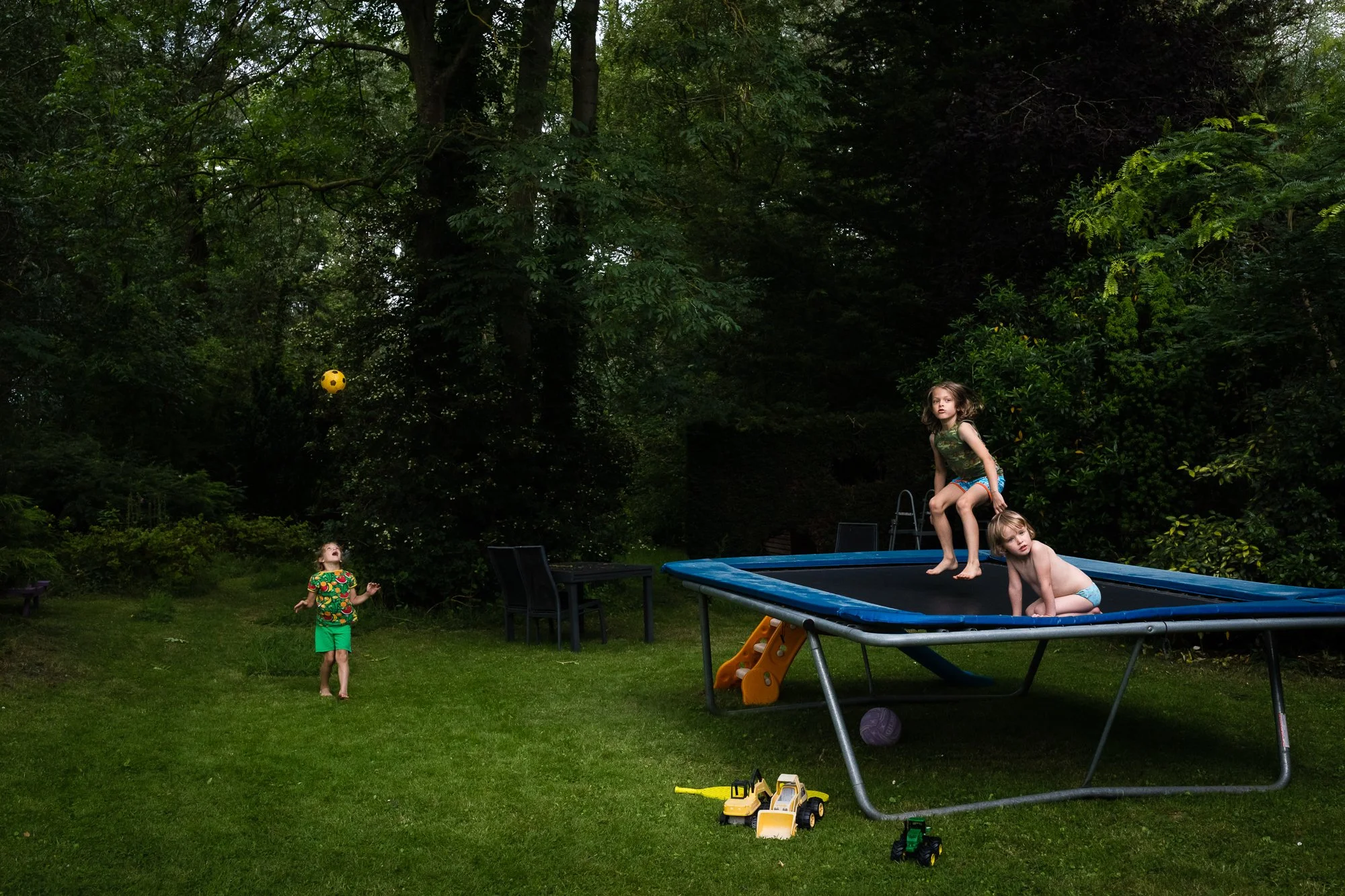 Three children playing in a backyard with a trampoline. One girl is on the trampoline, one girl is bouncing, and a boy is standing on the grass thrown a yellow ball. Toys are scattered on the grass, and there are green trees in the background.