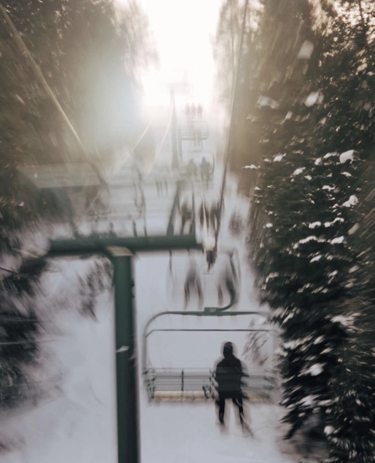 A person standing on a ski lift chair in a foggy, snowy landscape with trees and snow-covered ground.