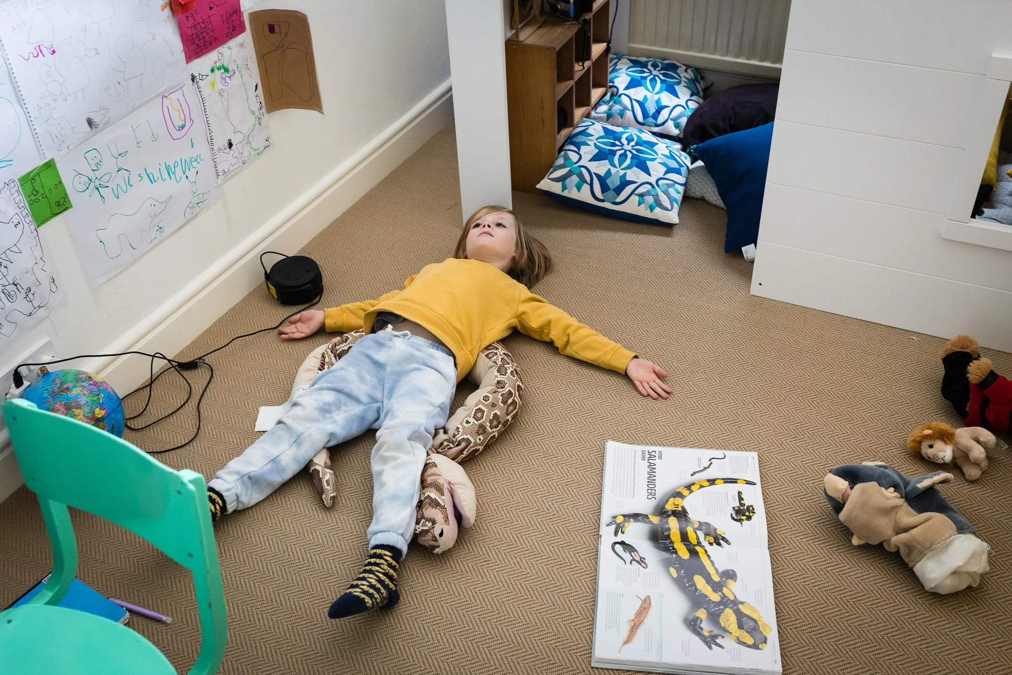 Child lying on the floor with arms and legs outstretched in a child’s bedroom, surrounded by toys, books, and artwork on the wall.