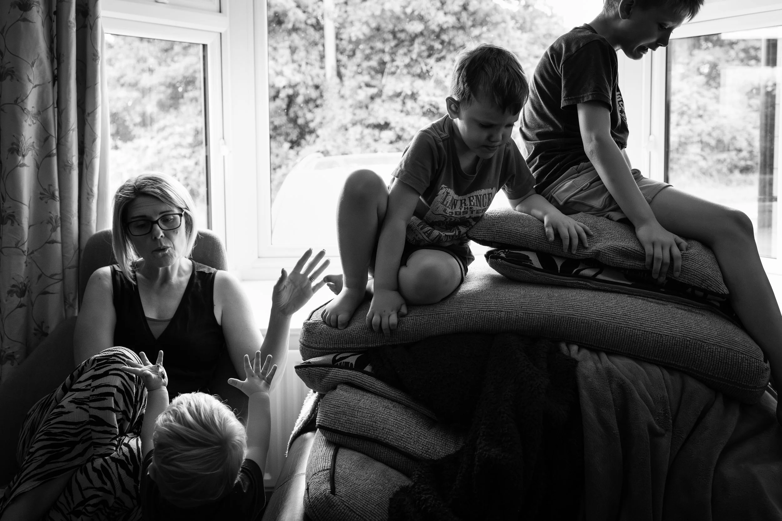 A woman sits on a couch with two children on a pile of cushions as they climb or jump, and a young child with arms raised lies on the floor nearby, inside a room with a large window.