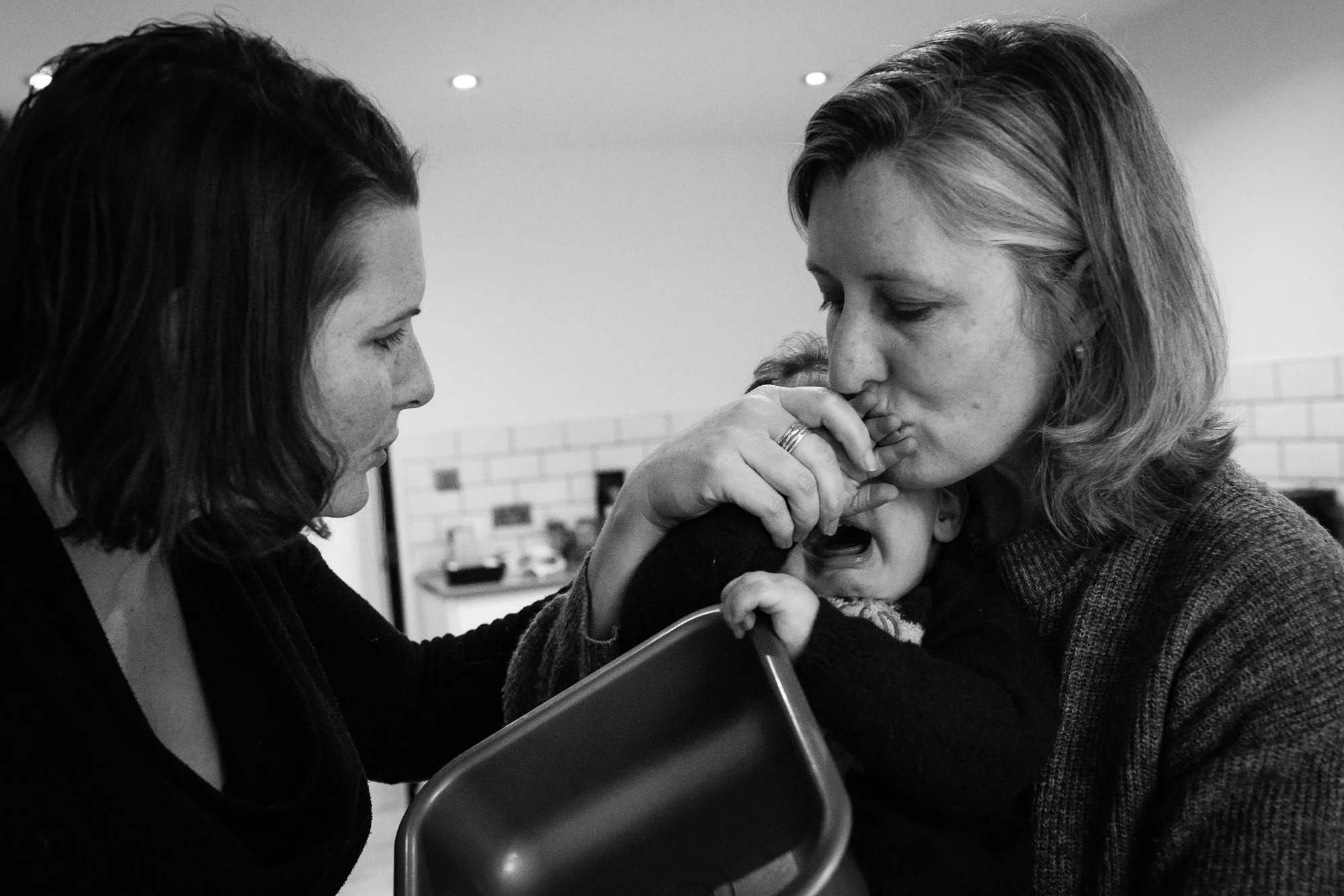 Two women comfort a crying baby, one woman holding the child's face and the other leaning in with concern, in an indoor setting.
