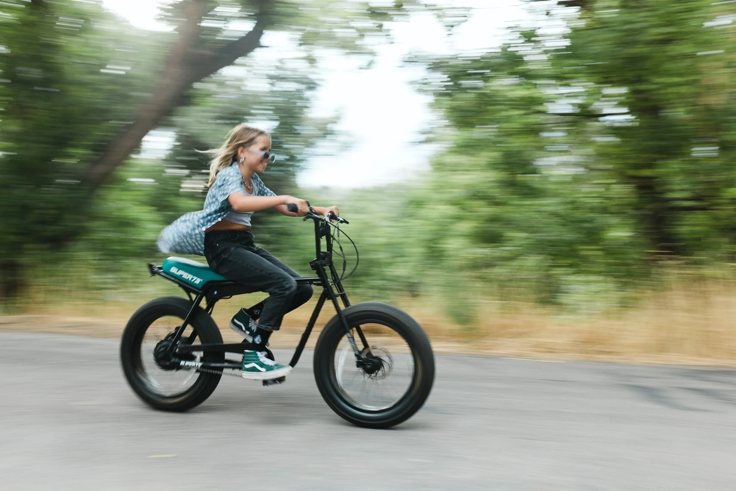 Woman riding an electric bicycle on a paved path with trees in the background, motion blur indicating speed.