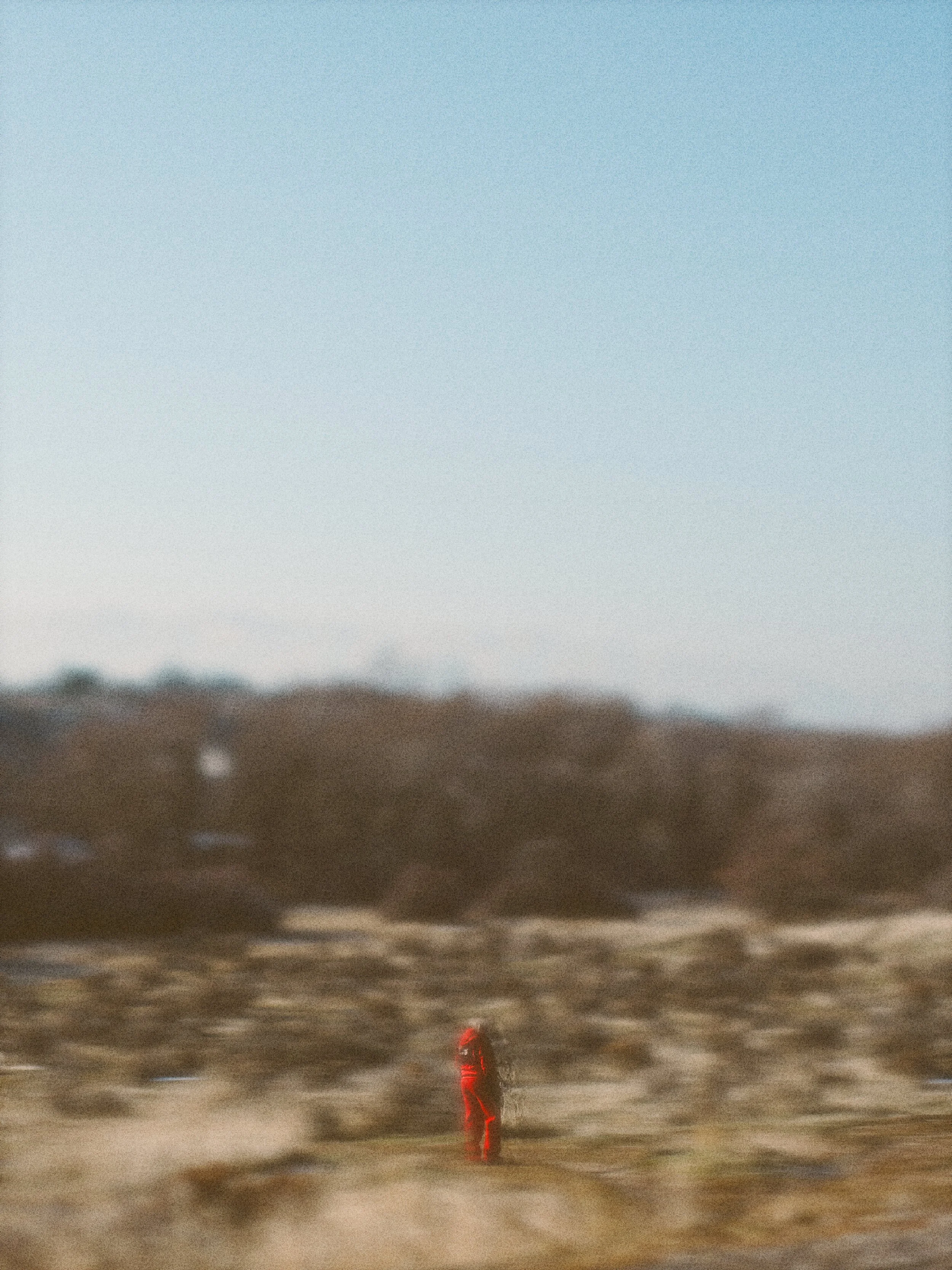 A person dressed in red walking in a barren, dusty landscape with hills and a clear sky.