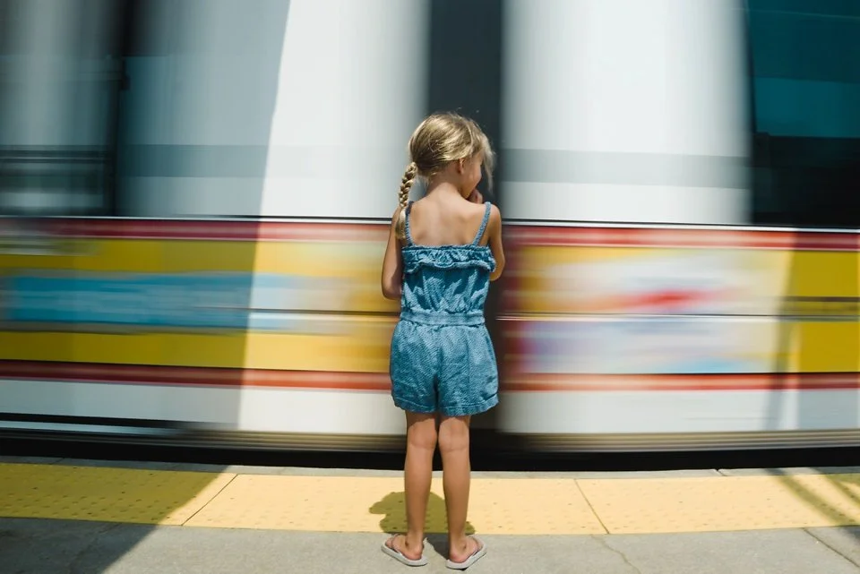 A young girl with blonde hair in a braid, wearing a blue romper, standing on a yellow tactile paving strip, watching a moving train at a station.