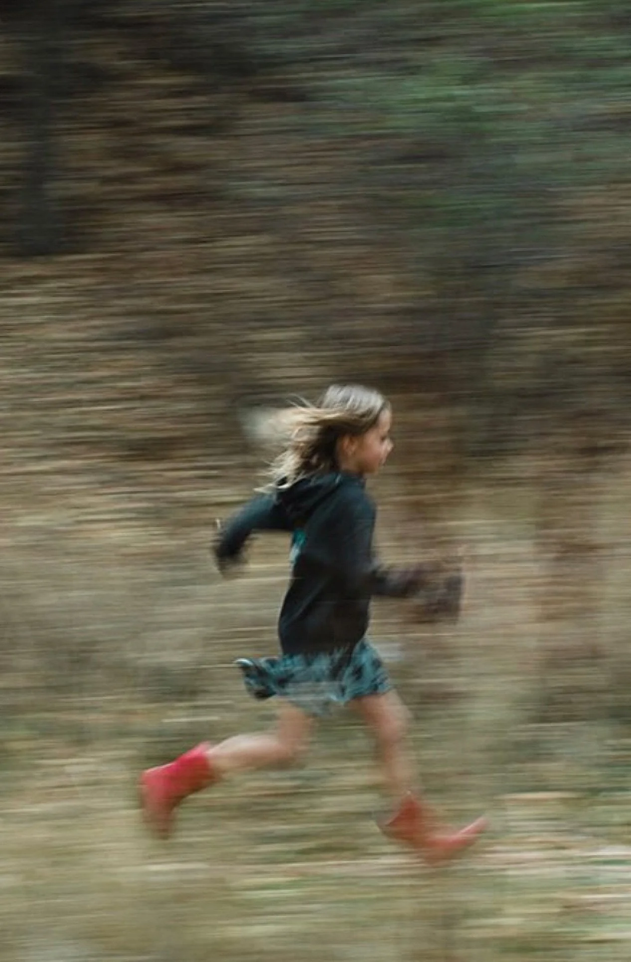 A young girl running outdoors through a wooded area, wearing a black jacket, patterned shorts, and red rain boots.